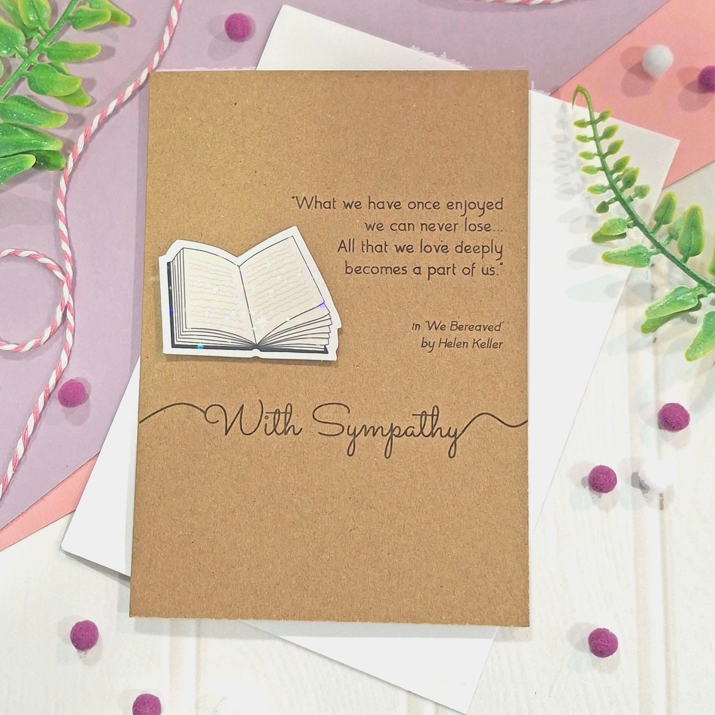 A sympathy card made from kraft card with a printed quote by Helen Keller from her book 'We Bereaved', a raised, holographic, illustrated open book embellishment, and the words 'With Sympathy' in elegant script, all styled on top of white and pink envelopes with decorative greenery and pom-poms.