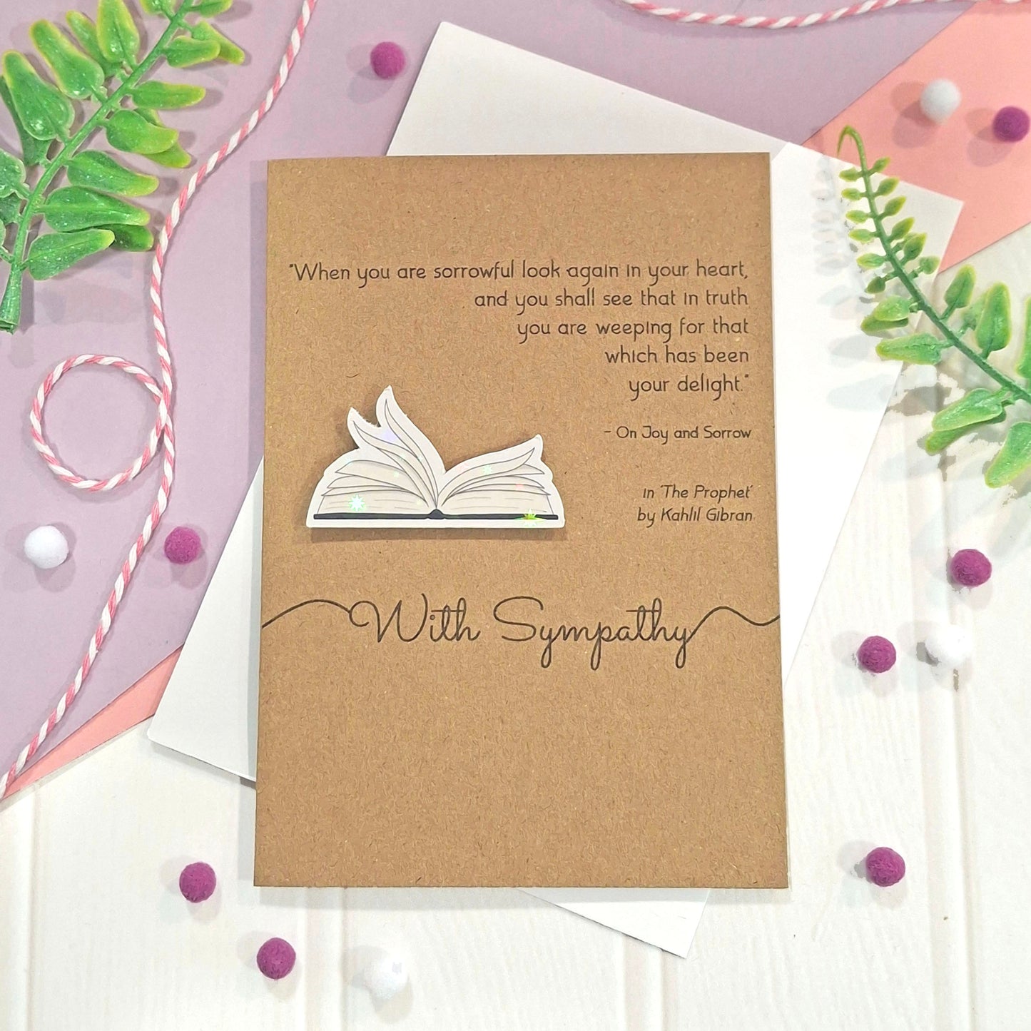 A kraft brown sympathy card featuring illustrated book pages in a raised, holographic embellishment, a printed quote from The Prophet by Kahlil Gibran, and the words With Sympathy in elegant black font. The card is styled in a flat-lay arrangement with a white envelope, pastel decorative leaves, and small purple and white pom-poms.