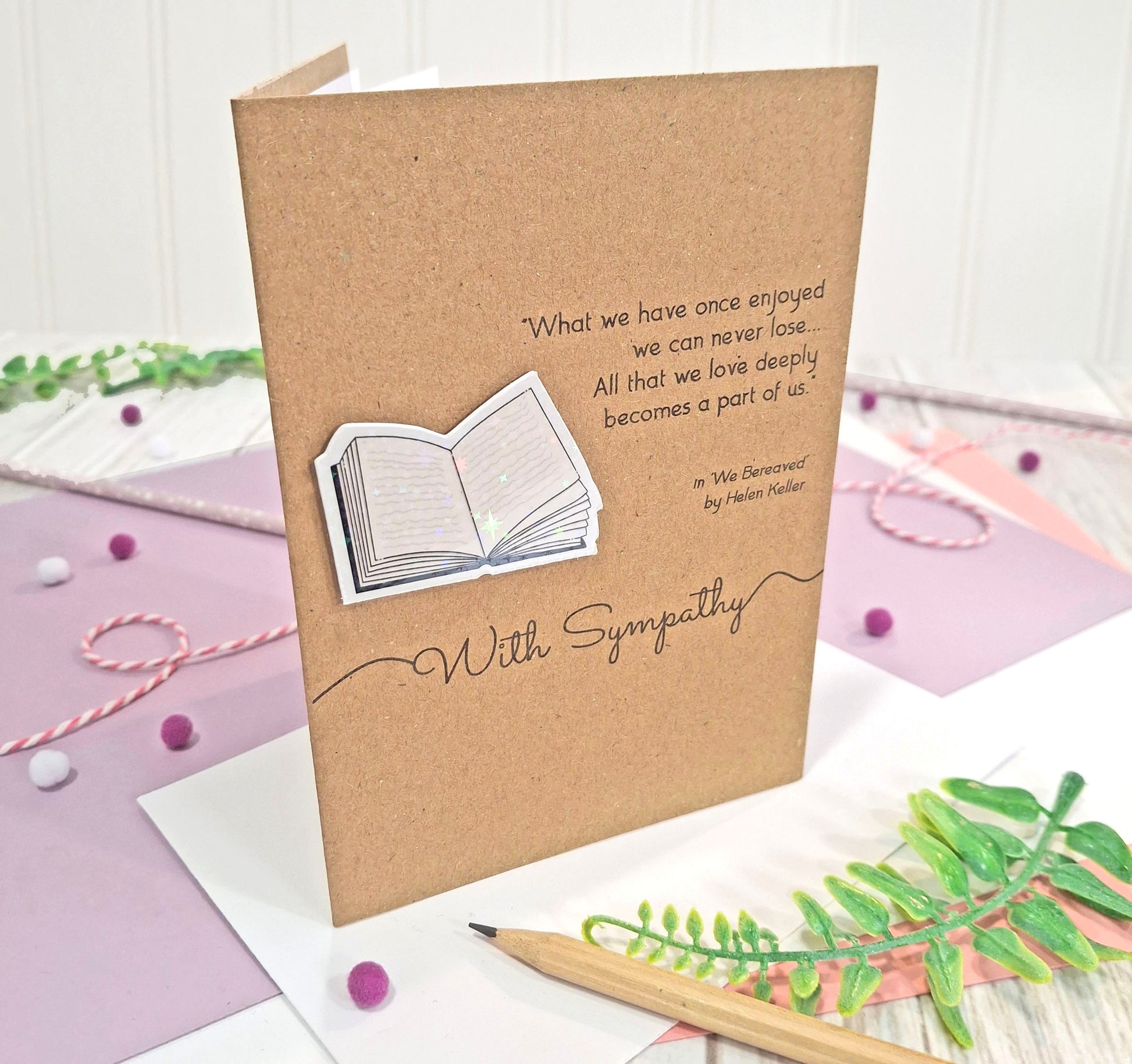 A sympathy card made from brown kraft card with a raised, holographic, illustrated open book embellishment, a printed quote from the book 'We Bereaved' by Helen Keller, and the words With Sympathy.