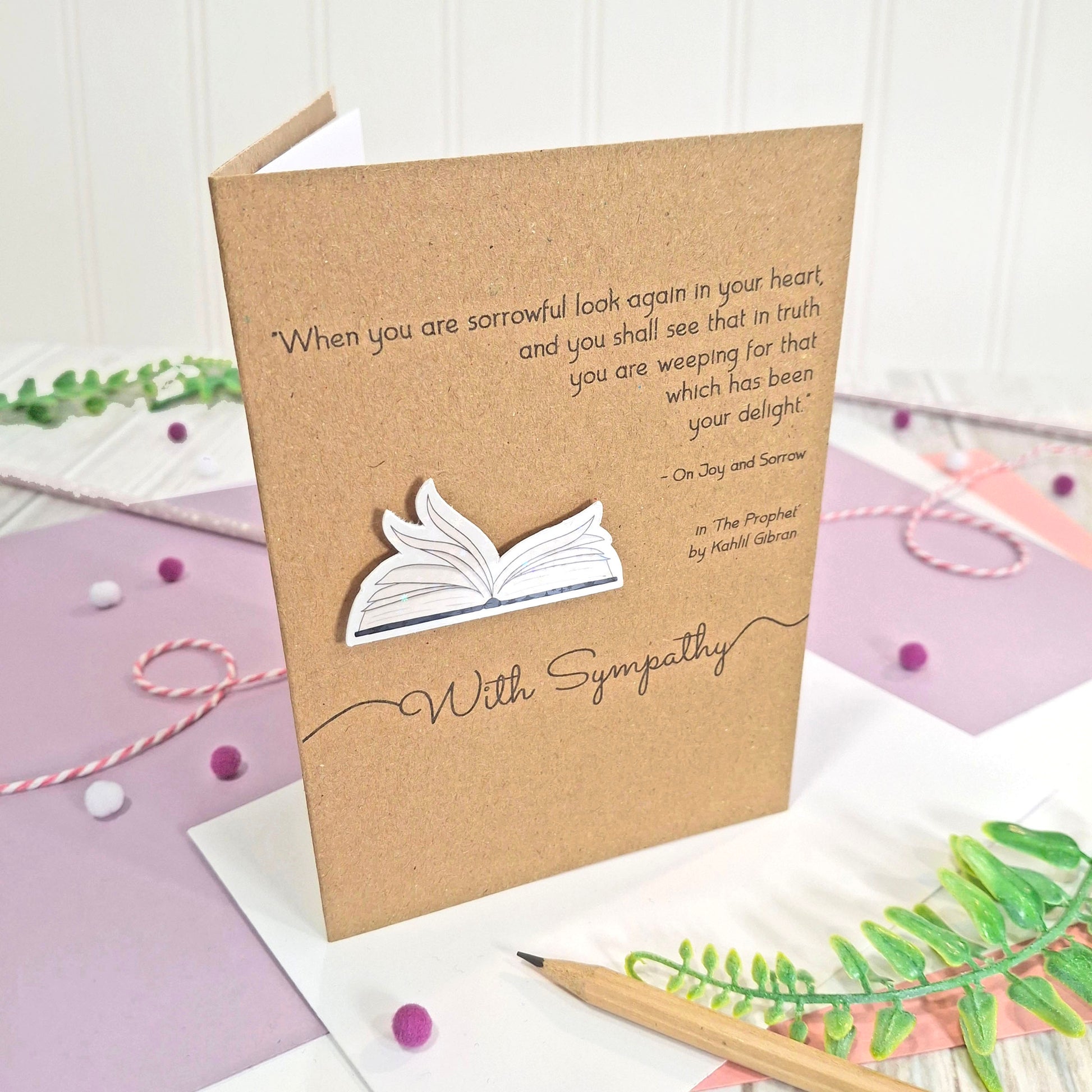 A sympathy card made from brown kraft card with a printed quote by Kahlil Gibran from The Prophet, the words 'With Sympathy' and illustrated book pages in a raised, holographic embellishment, all displayed on a styled pastel surface with string, paper, and greenery.
