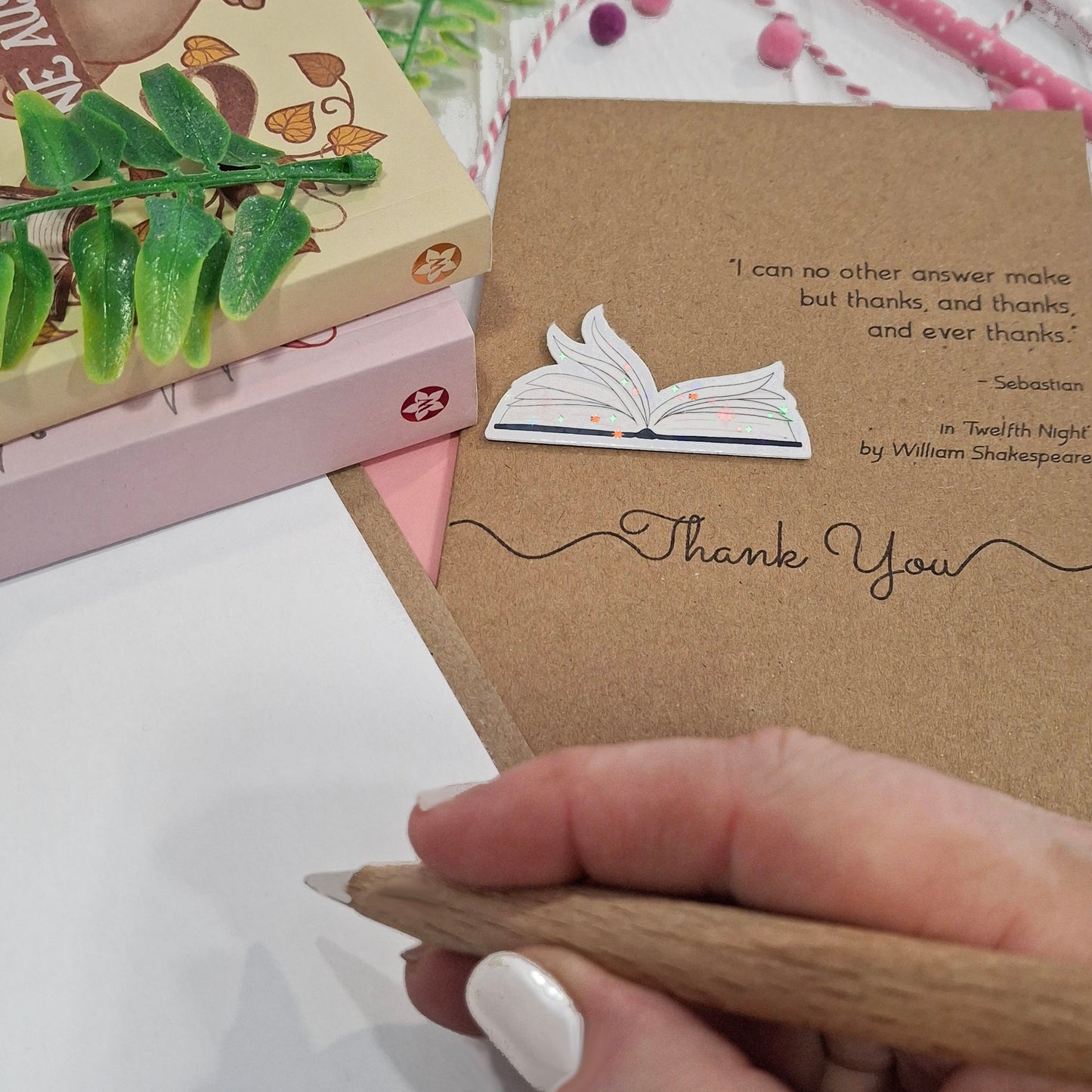 A close up of a hand holding a wooden pen, and writing inside a card. The card features a raised holographic embellishment of an open book with gently fanned pages, the words Thank You, and a quote from Twelfth Night by William Shakespeare.
