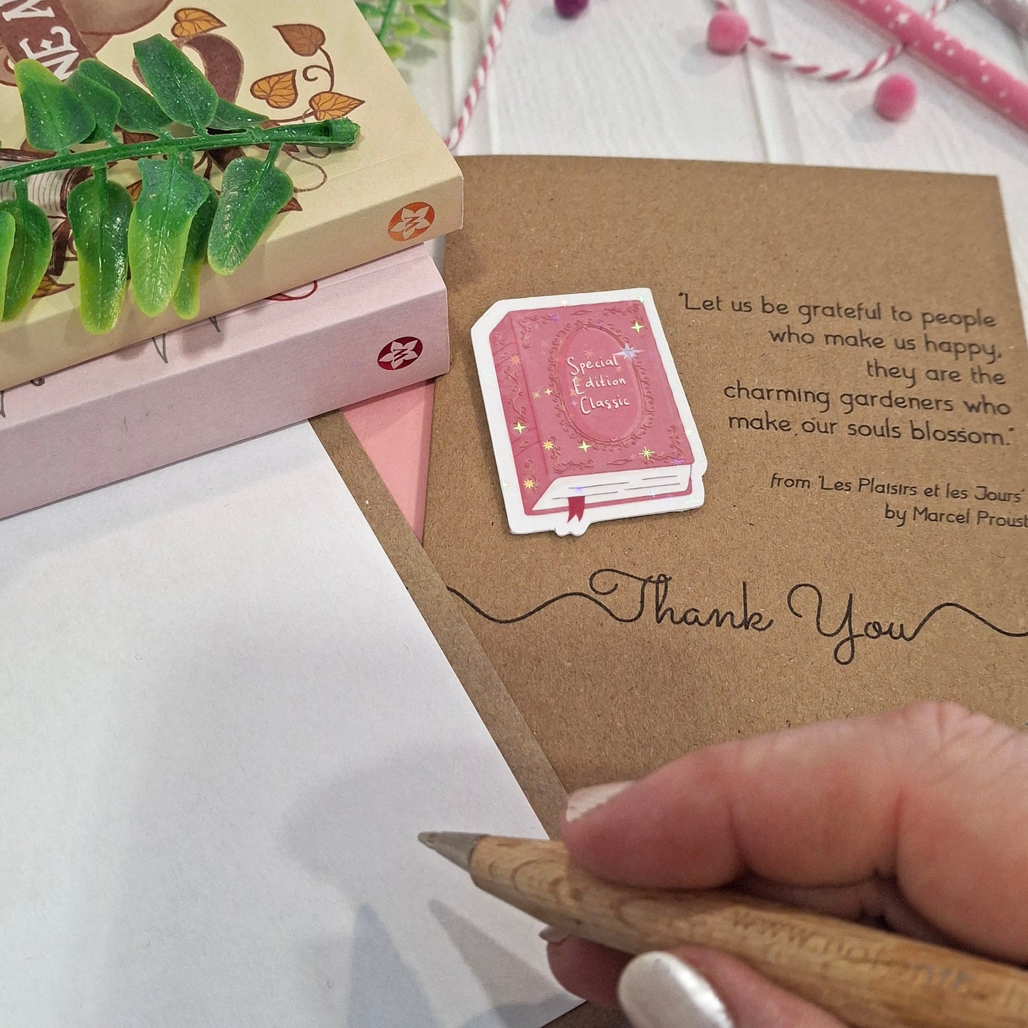 A close-up of a hand holding a wooden pen and writing inside a card. The card features a raised holographic embellishment of a pink special edition classic book, the words Thank You, and a quote from Marcel Proust.