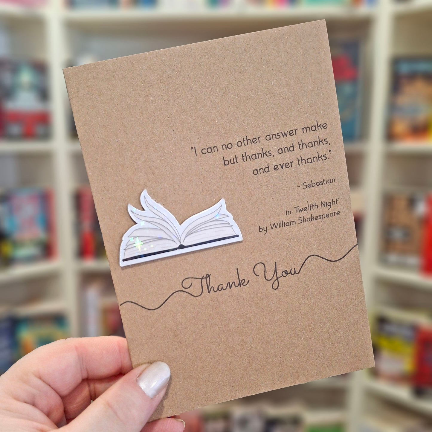 A hand holds a brown kraft thank you card featuring a raised holographic embellishment of an open book with gently fanned pages, and a printed quote by William Shakespeare. The background shows a blurred bookshop interior.
