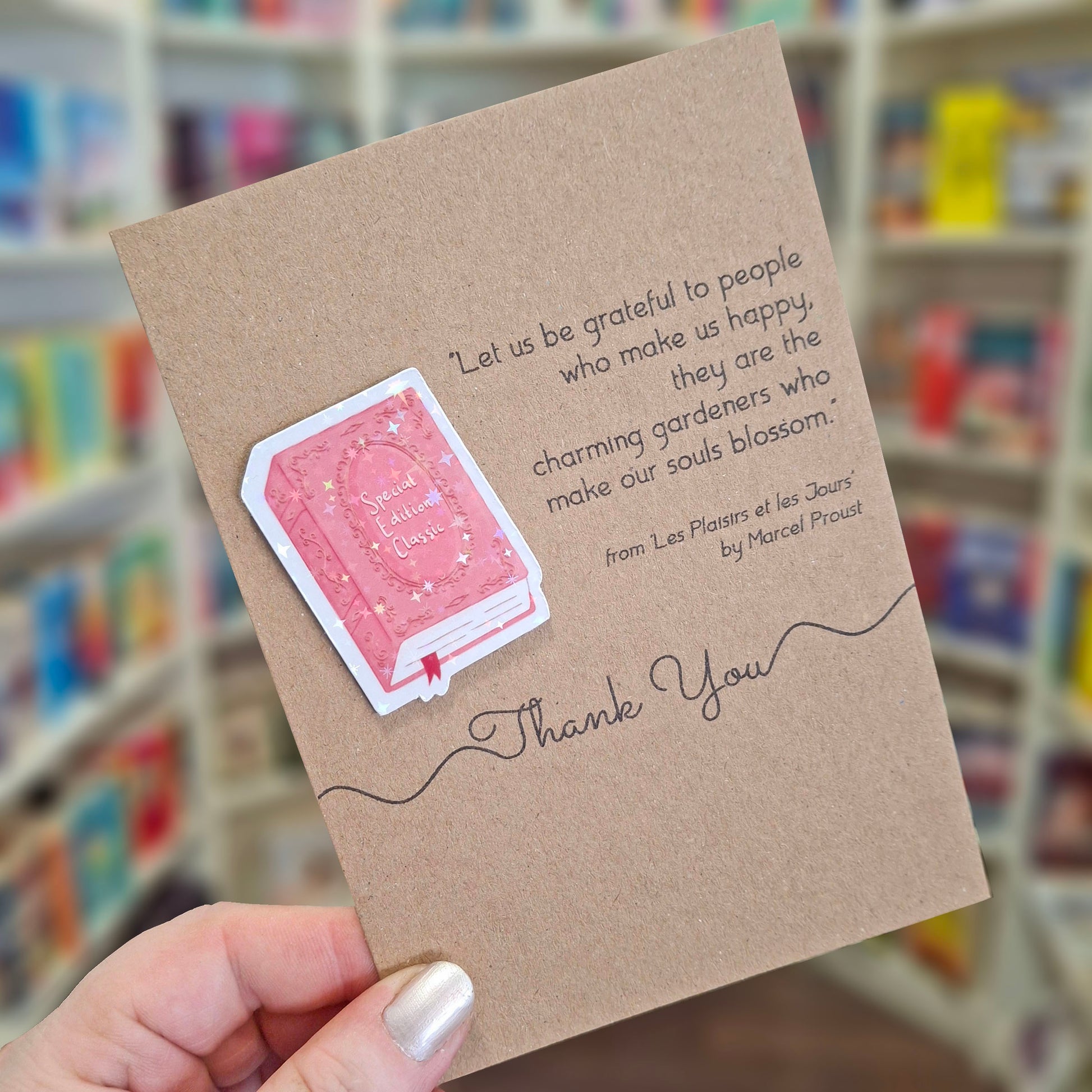 A hand holds a brown kraft thank you card featuring a raised holographic embellishment of a pink special edition classic book, and a printed quote by Marcel Proust. The background shows a blurred bookshop interior.