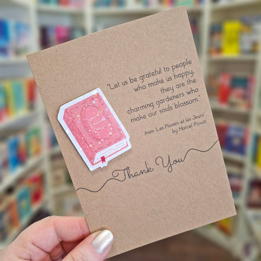 A hand holds a brown kraft thank you card featuring a raised holographic embellishment of a pink special edition classic book, and a printed quote by Marcel Proust. The background shows a blurred bookshop interior.