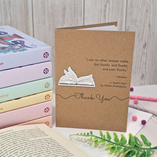 A brown kraft thank you card styled on a tabletop with books, an open book, and greenery. The card features a raised holographic embellishment of an open book with gently fanned pages, the words Thank You, and a quote from Twelfth Night by William Shakespeare.