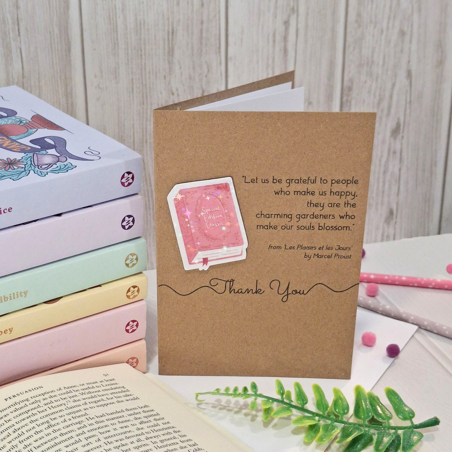A brown kraft thank you card, styled on a desk with books, an open book, and a green sprig. The card features a raised holographic embellishment of a pink special edition classic book, the words Thank You, and a quote from Marcel Proust.