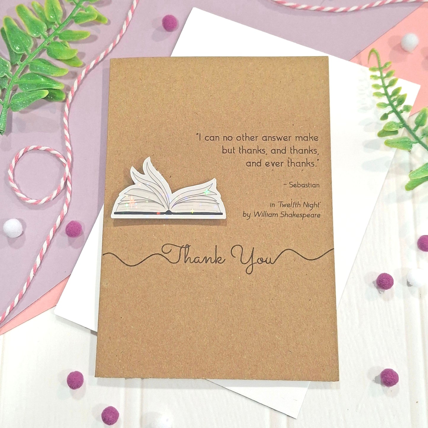 A kraft thank you card featuring illustrated book pages in a raised, holographic embellishment, a printed quote from William Shakespeare, and the words Thank You, all styled on top of white envelopes with decorative twine, green leaves, and purple pom-poms.