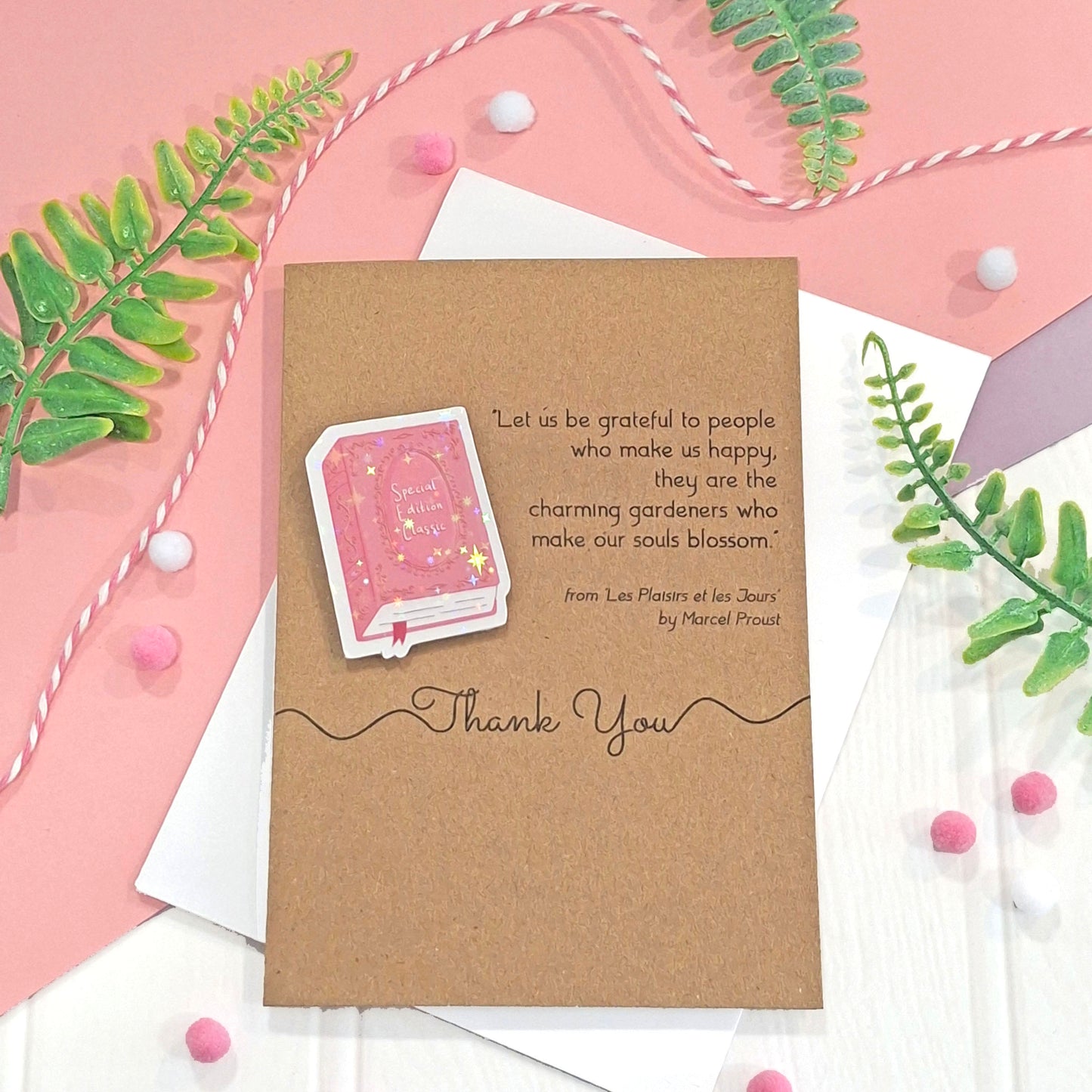 A brown kraft thank you card with a raised, holographic, illustrated story book embellishment in pink, a quote by Marcel Proust, and the words 'Then You' in black script, all styled in a flat lay on a pink and white background with decorative green leaves and pom-poms.