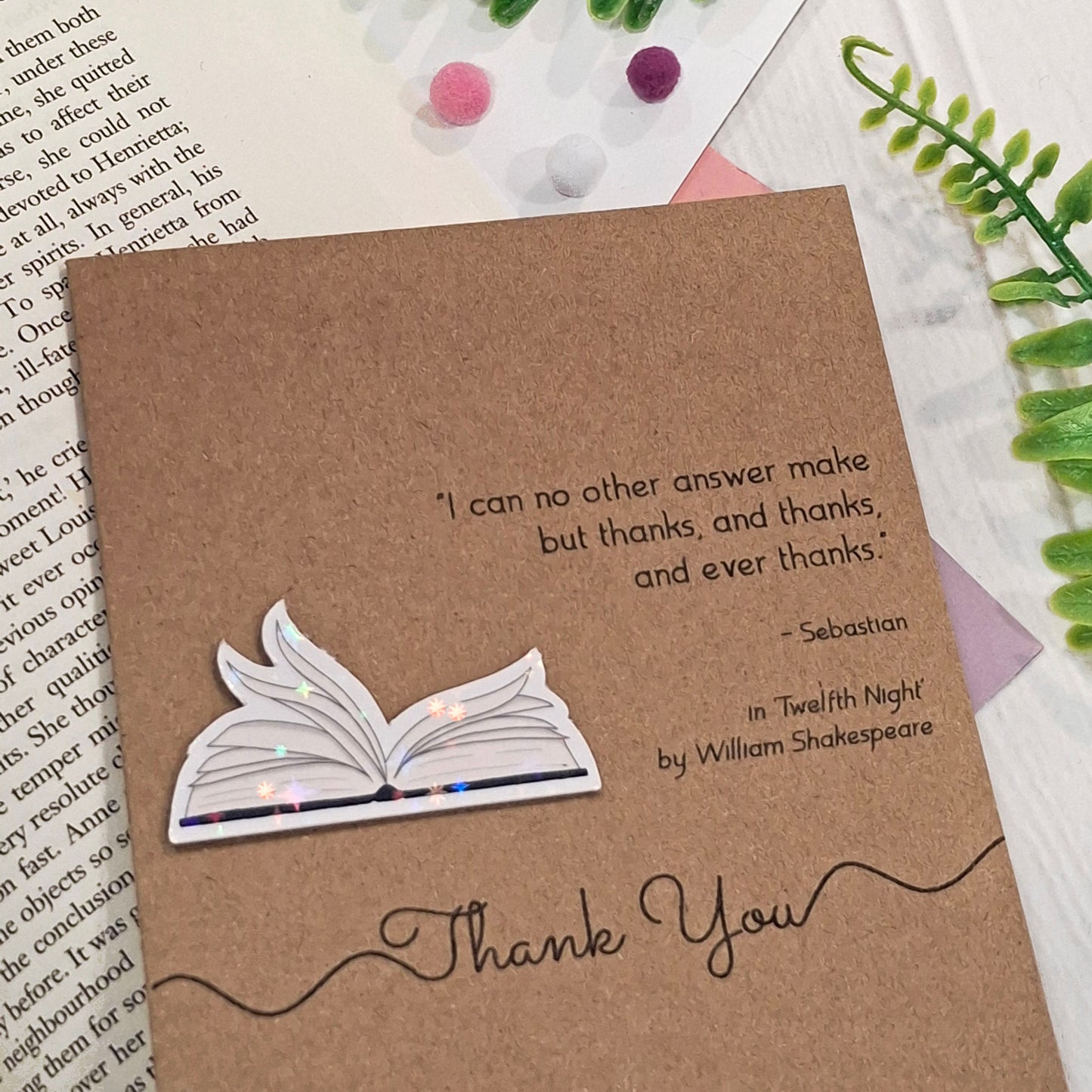 A brown kraft thank you card placed on a white surface with decorative green leaves and small pom-poms. The card features a raised holographic embellishment of an open book with gently fanned pages, the words Thank You, and a quote from Twelfth Night by William Shakespeare.