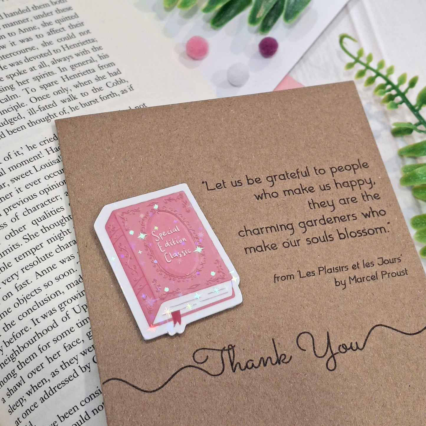 A brown kraft thank you card styled on a book page with small decorative pom-poms and greenery. The card features a raised holographic embellishment of a pink special edition classic book, the words Thank You, and a quote from Marcel Proust.