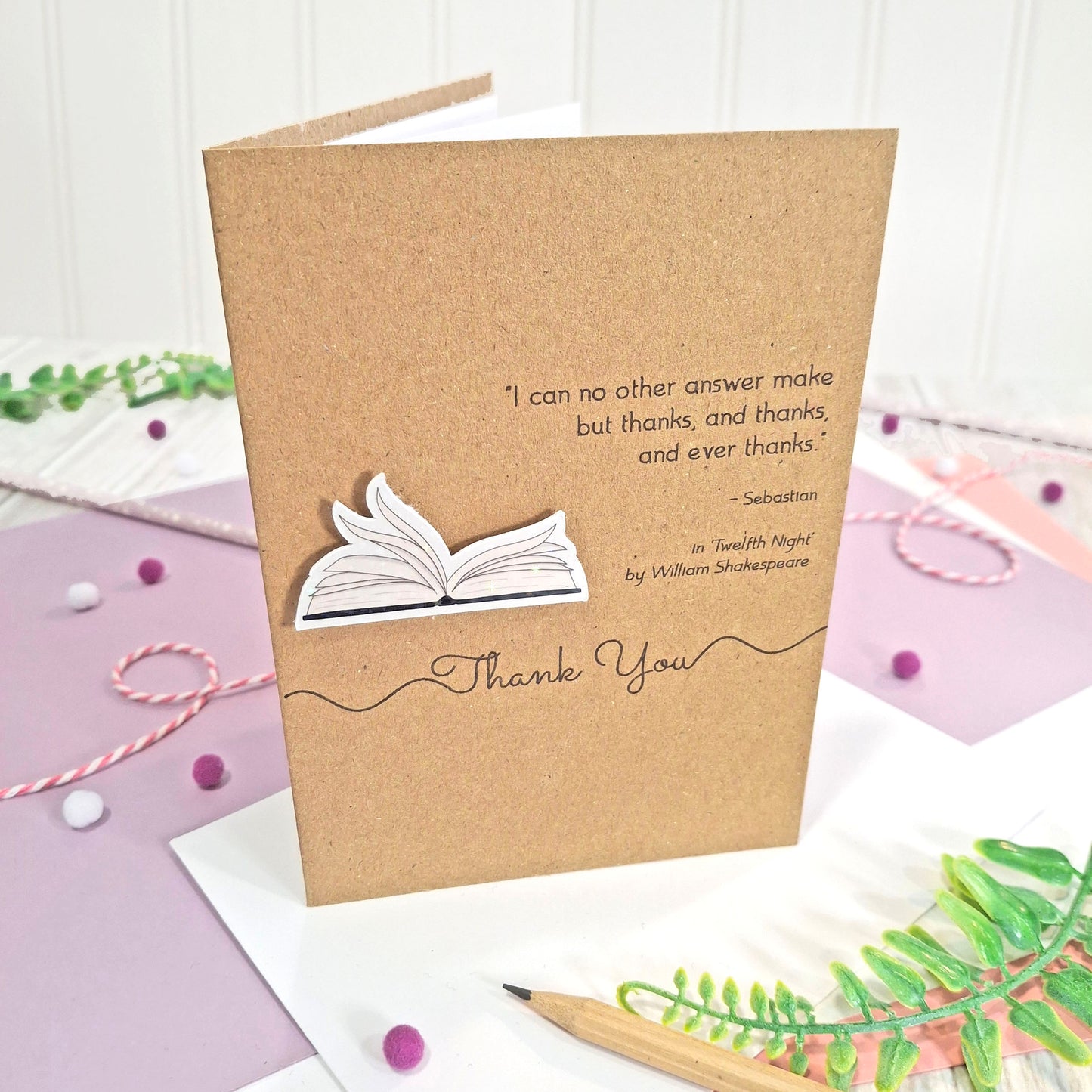 A kraft thank you card with illustrated book pages in a raised, holographic embellishment, the words Thank You, and a quote from William Shakespeare's Twelfth Night, all displayed on a styled flat-lay surface with pastel paper, a pencil, and decorative elements.