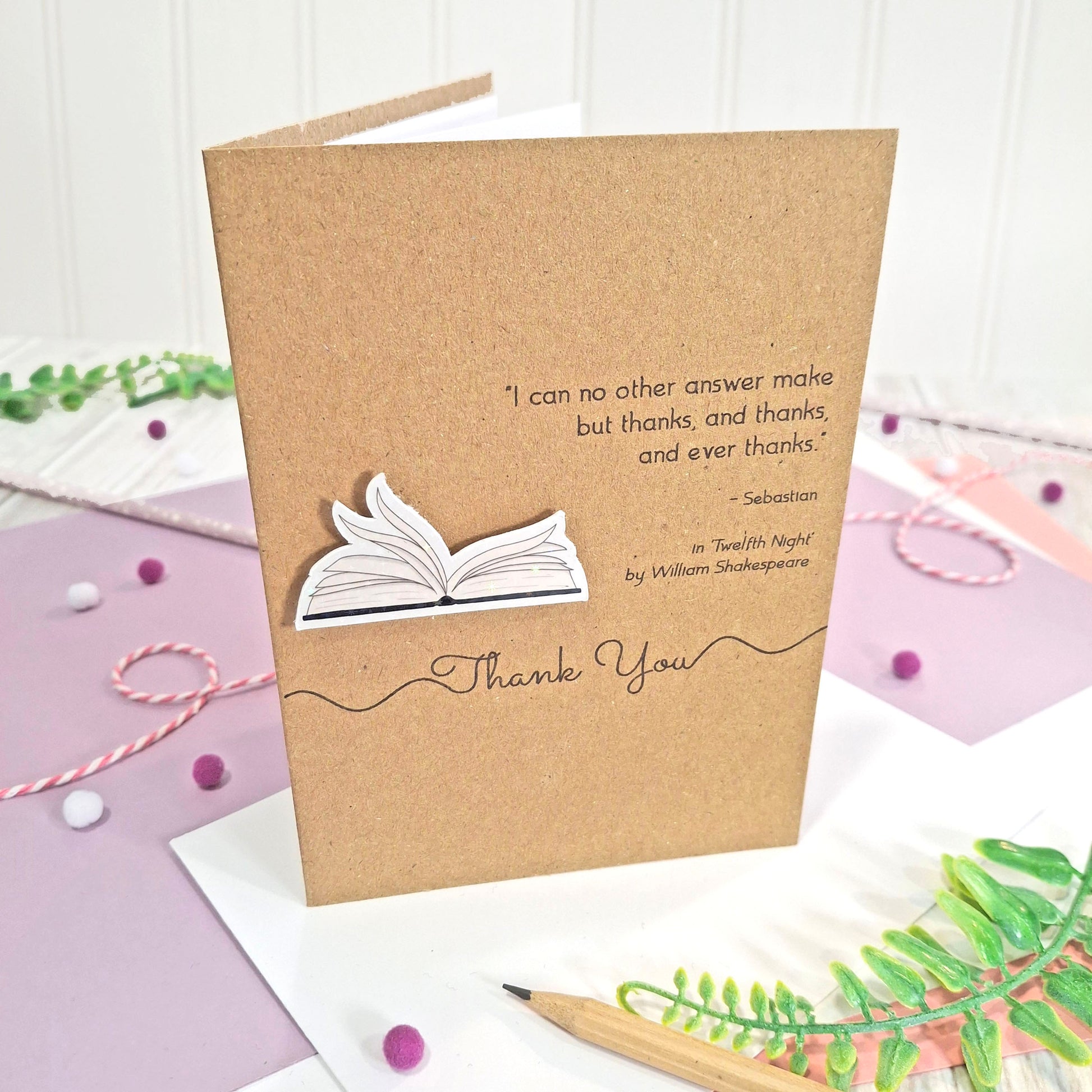 A kraft thank you card with illustrated book pages in a raised, holographic embellishment, the words Thank You, and a quote from William Shakespeare's Twelfth Night, all displayed on a styled flat-lay surface with pastel paper, a pencil, and decorative elements.