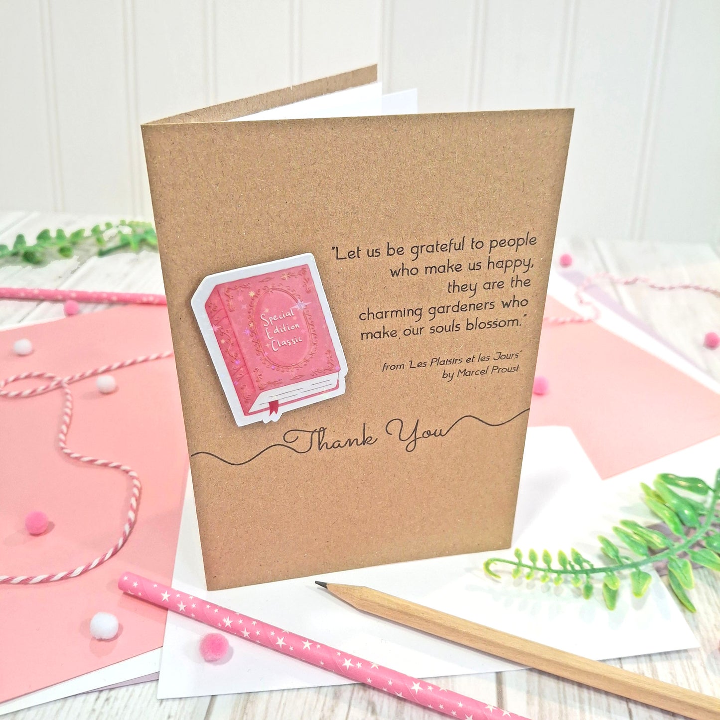 A brown kraft thank you card standing upright on a styled desk with a raised, holographic, illustrated story book embellishment in pink, a quote by Marcel Proust, and the words Thank You. The card is styled on a background of pink paper, with a pencil, and greenery.