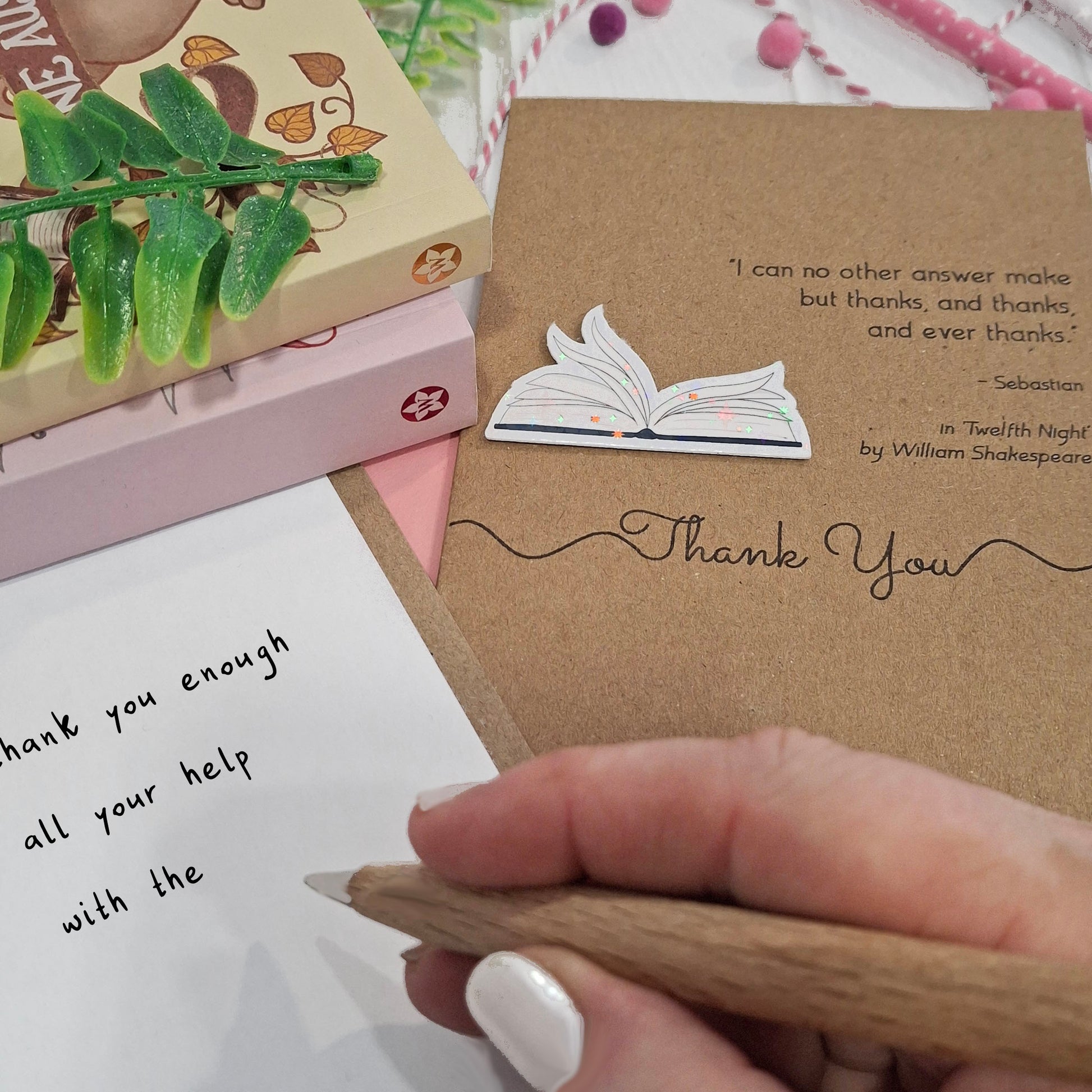 A close up of a hand holding a wooden pen, and writing a thank you message inside a card. The card features a raised holographic embellishment of an open book with gently fanned pages, the words Thank You, and a quote from Twelfth Night by William Shakespeare.