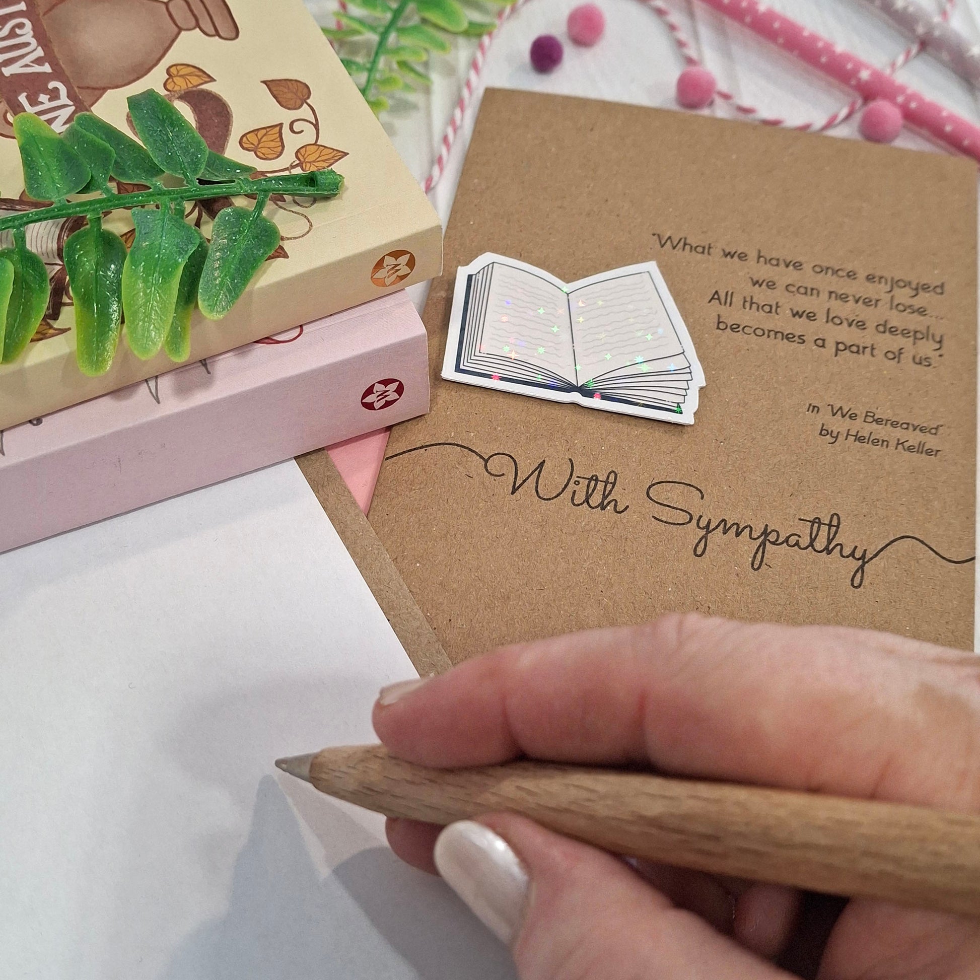 A close-up of a hand holding a wooden pen and writing inside a card. The card features a raised holographic embellishment of an open book, and reads 'With Sympathy', with a quote from We Bereaved by Helen Keller. 