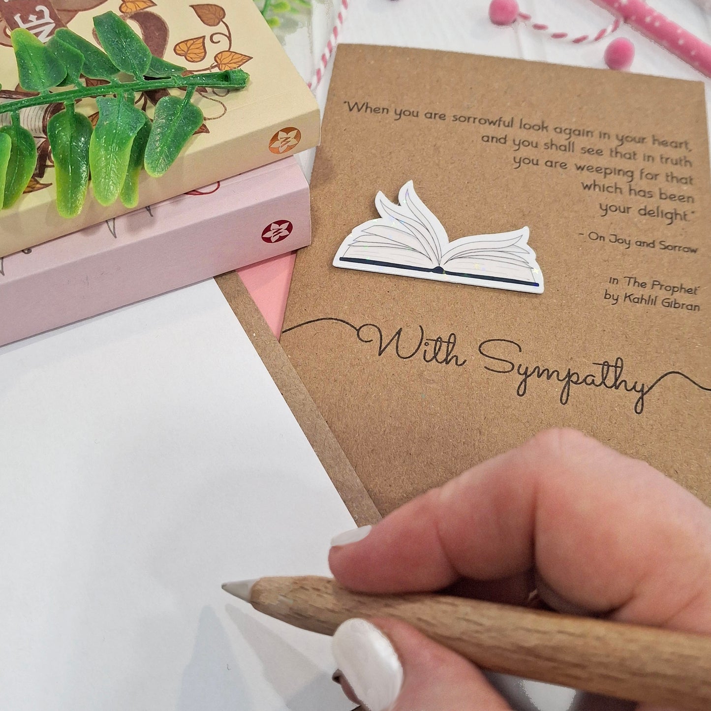 A close up of a hand holding a wooden pen, and writing inside a card. The card features a raised holographic embellishment of an open book with gently fanned pages, the words With Sympathy, and a quote from The Prophet by Kahlil Gibran.