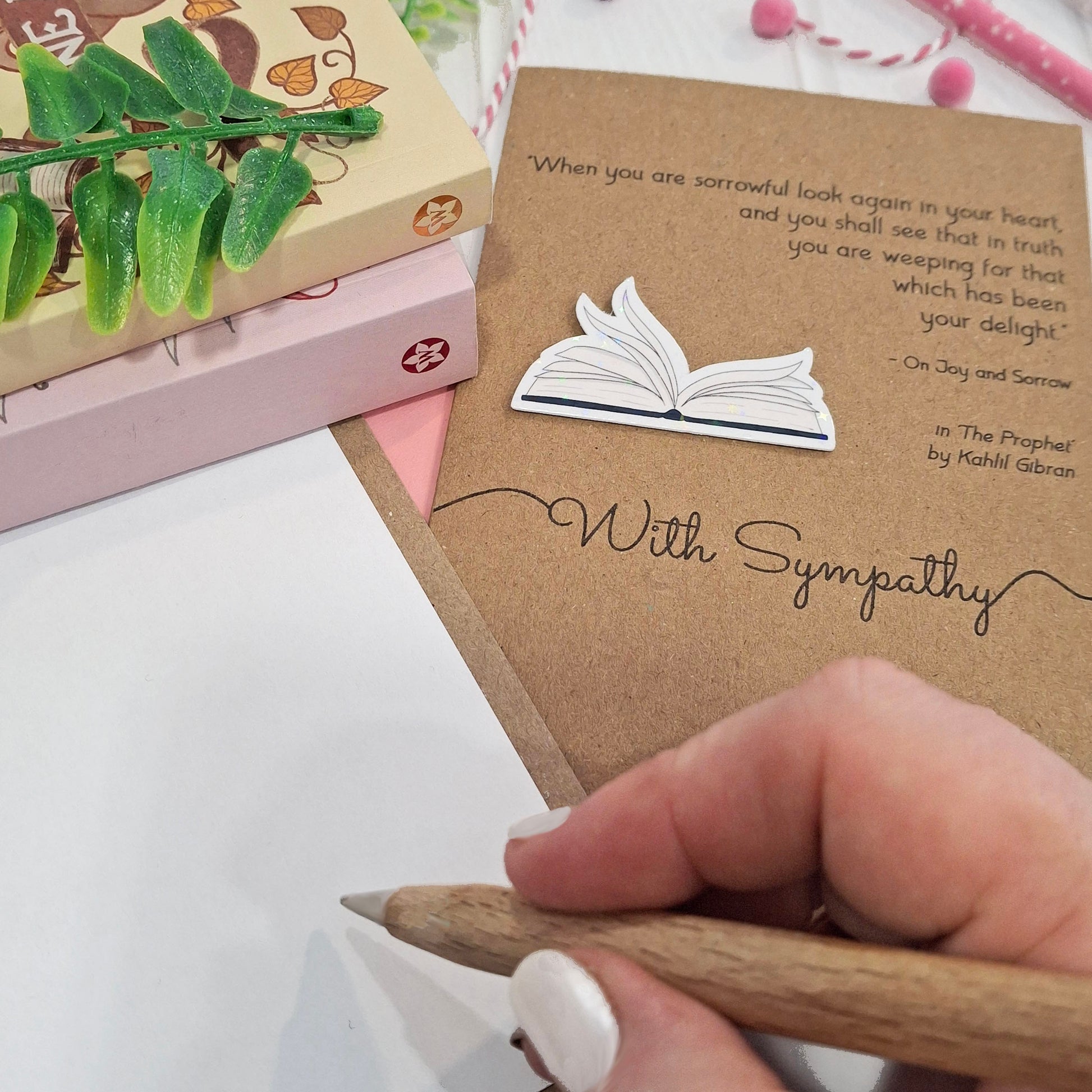 A close up of a hand holding a wooden pen, and writing inside a card. The card features a raised holographic embellishment of an open book with gently fanned pages, the words With Sympathy, and a quote from The Prophet by Kahlil Gibran.