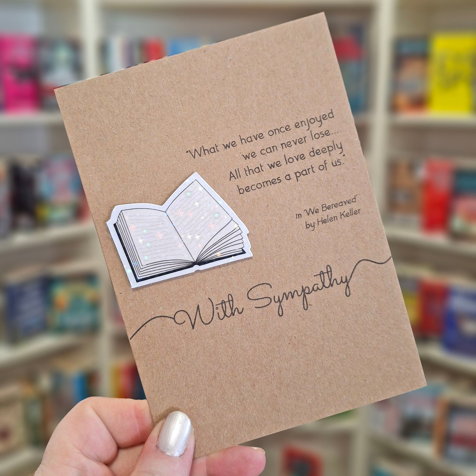 A hand holds a brown kraft sympathy card featuring a raised holographic embellishment of an open book, and a printed quote by Helen Keller. The background shows a blurred bookshop interior.
