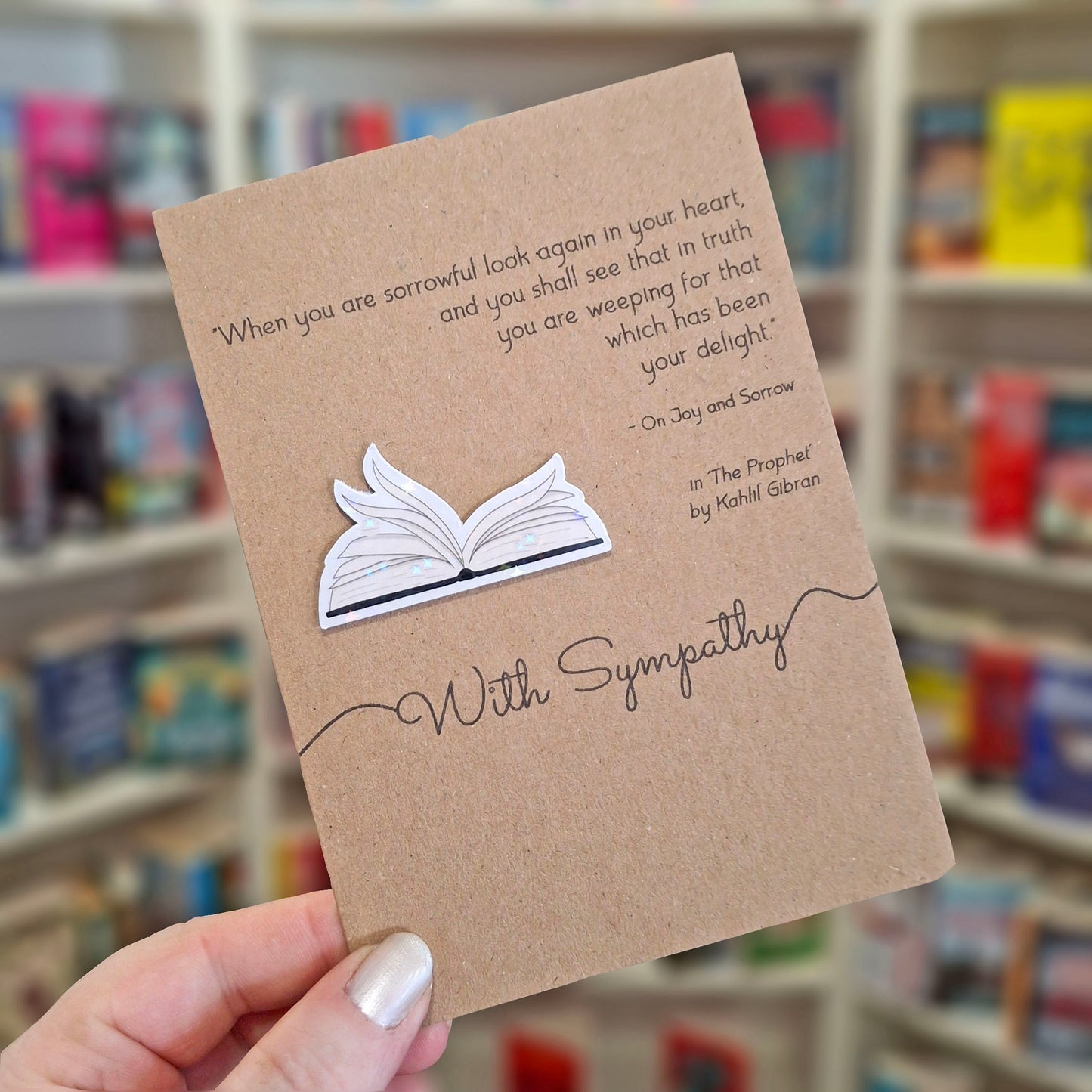 A hand holds a brown kraft sympathy card featuring a raised holographic embellishment of an open book with gently fanned pages, and a printed quote by Kahlil Gibran. The background shows a blurred bookshop interior.