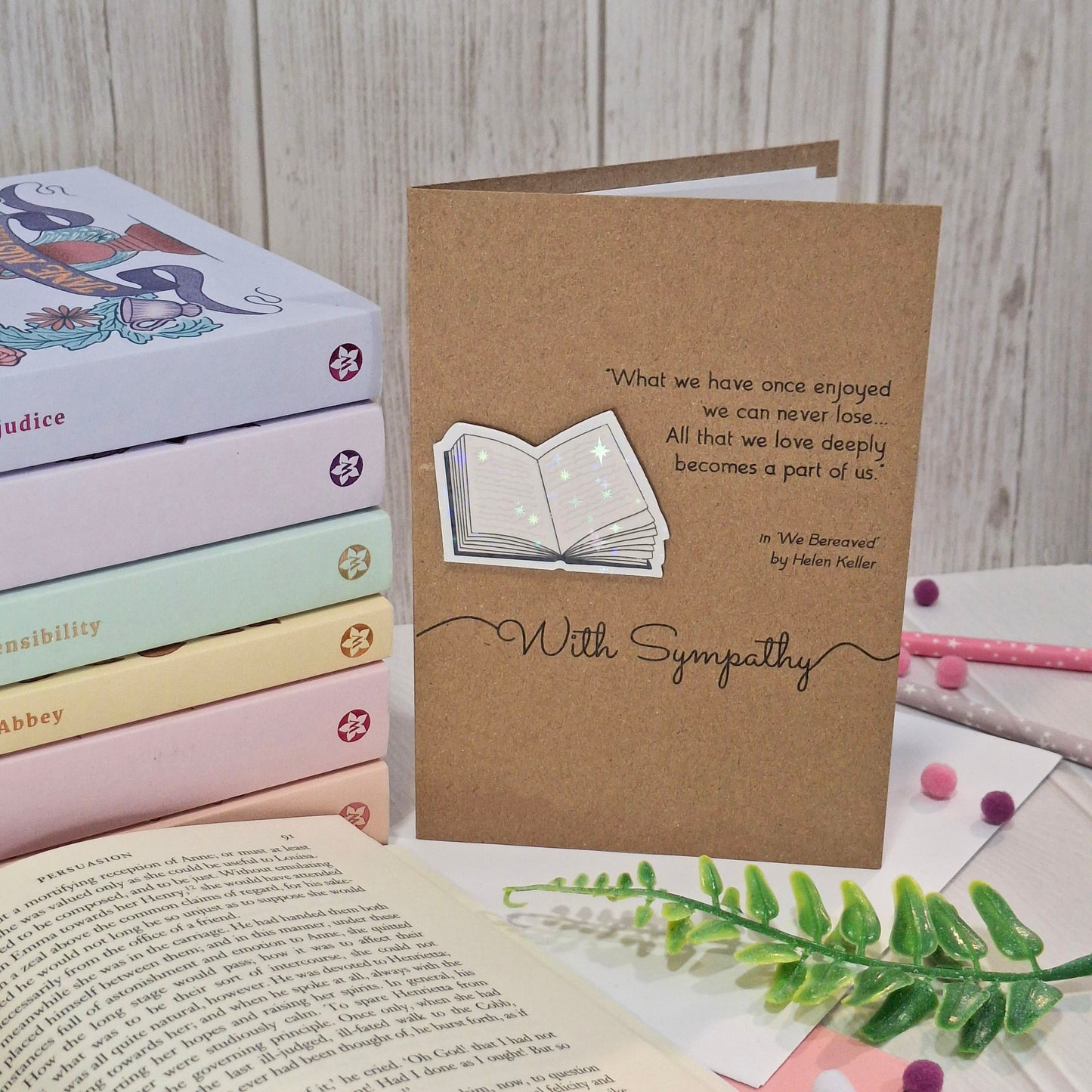 A brown kraft paper sympathy card, styled on a tabletop with pastel coloured books, and a green fern in the background. The card features a raised holographic embellishment of an open book, and reads 'With Sympathy', with a quote from We Bereaved by Helen Keller.