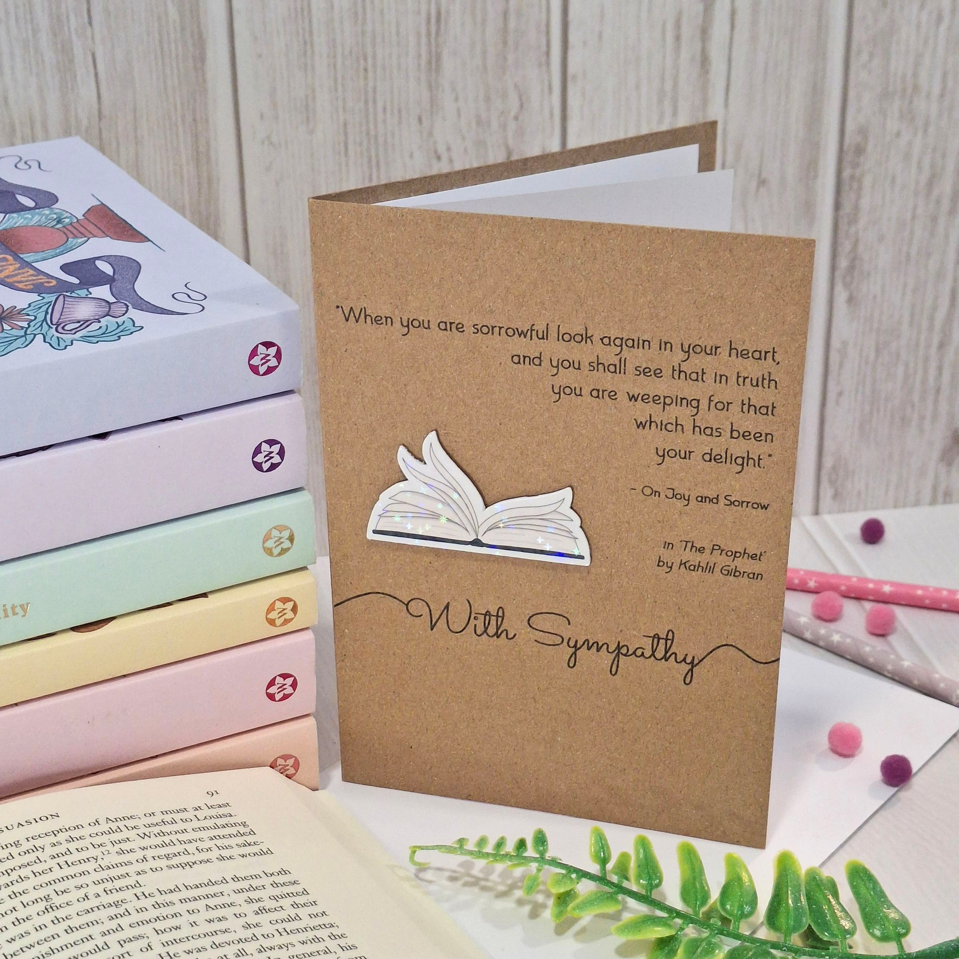 A sympathy card made from brown kraft card, standing upright on a tabletop with books, an open book, and greenery around it. The card features a raised holographic embellishment of an open book with gently fanned pages, the words With Sympathy, and a quote from The Prophet by Kahlil Gibran.