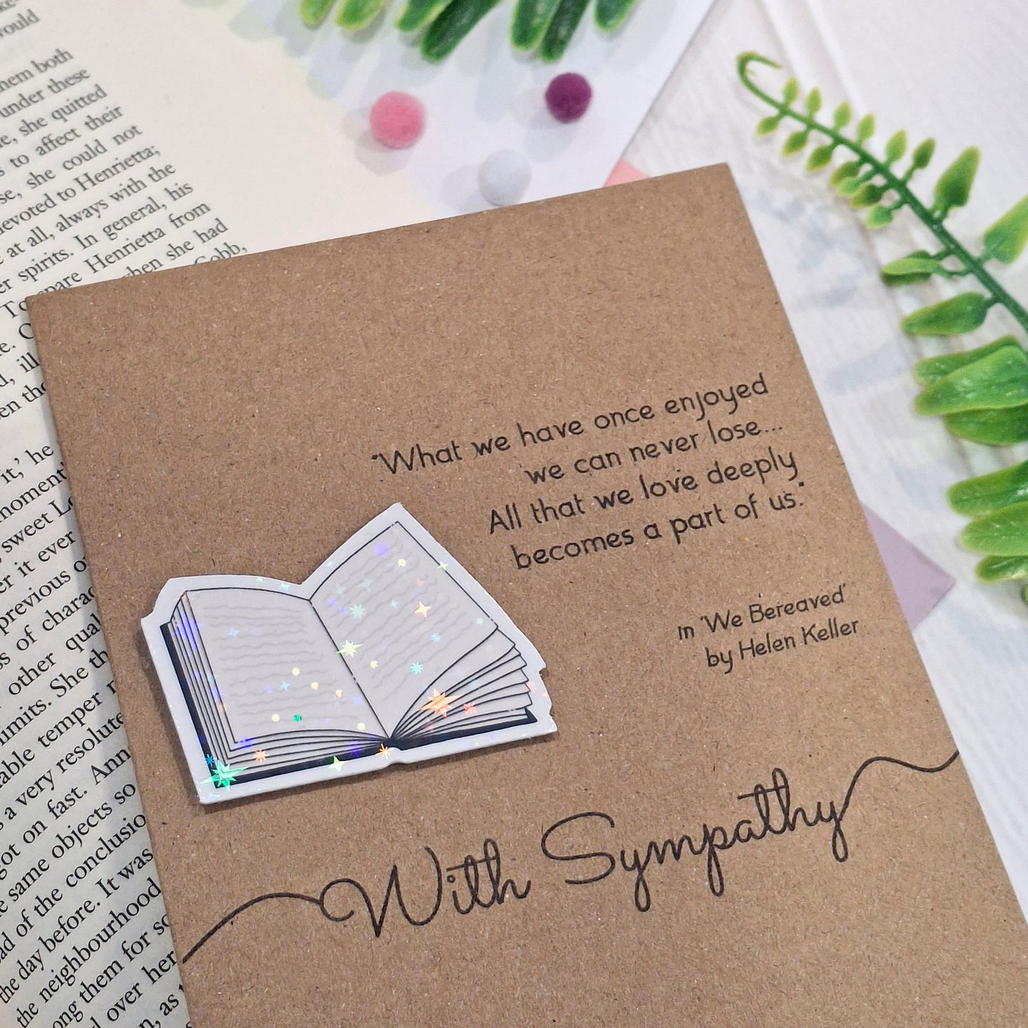 A sympathy card styled on top of an open book with decorative green leaves and small pom-poms. The card features a raised holographic embellishment of an open book, and reads 'With Sympathy', with a quote from We Bereaved by Helen Keller.