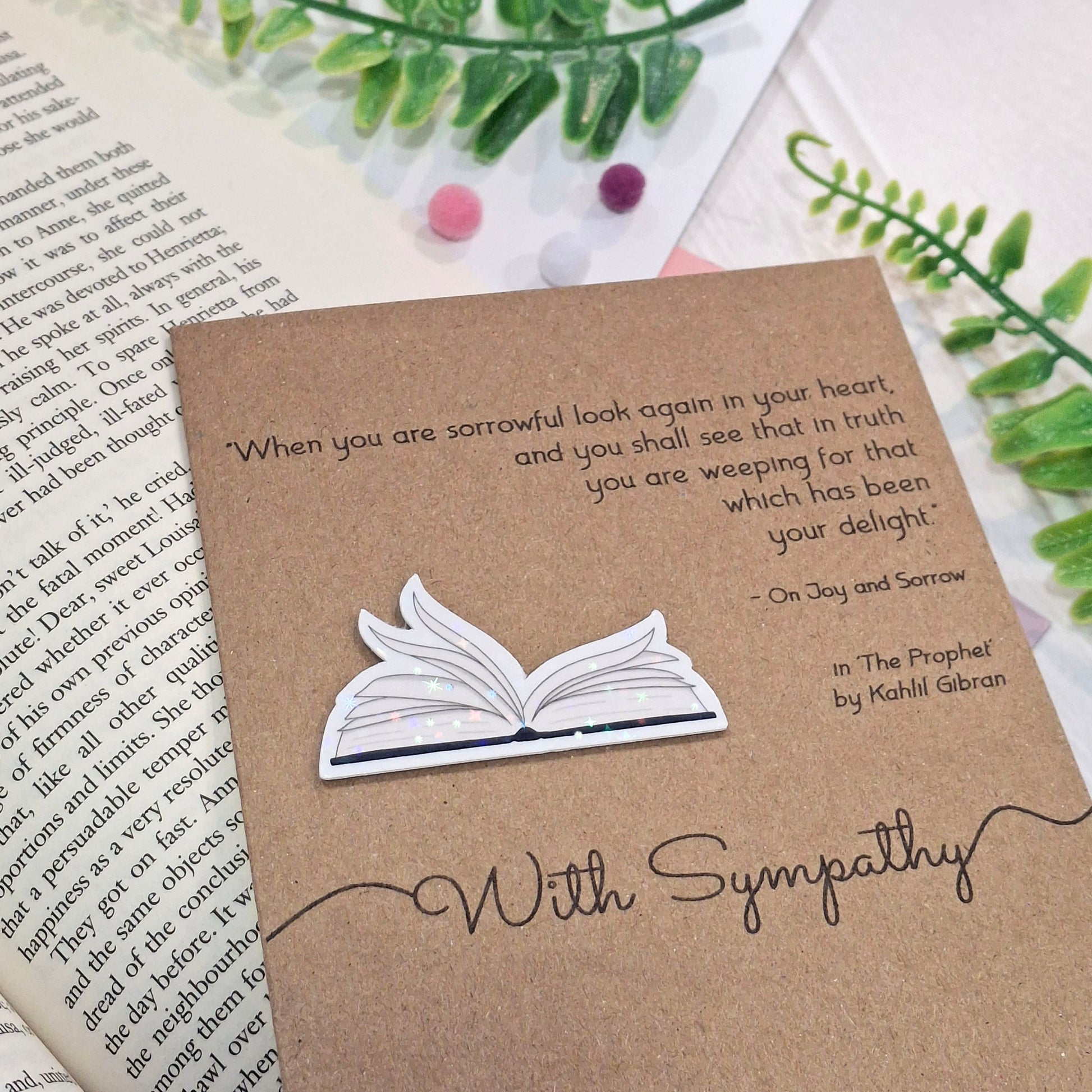 A sympathy card made from brown kraft card styled on top of an open book with green leaves and small pom-poms. The card features a raised holographic embellishment of an open book with gently fanned pages, the words With Sympathy, and a quote from The Prophet by Kahlil Gibran.