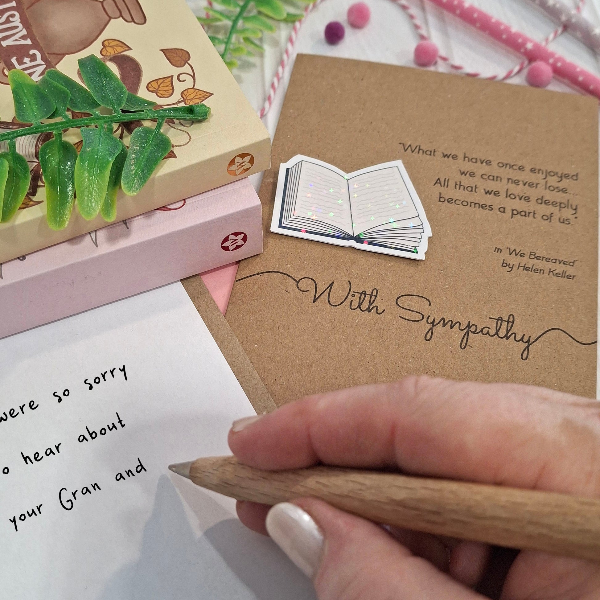 A close-up of a hand holding a wooden pen and writing a condolence message inside a card. The card features a raised holographic embellishment of an open book, and reads 'With Sympathy', with a quote from We Bereaved by Helen Keller. 