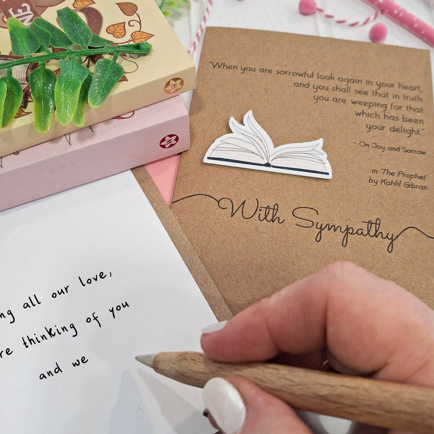 A close up of a hand holding a wooden pen, and writing a sympathy message inside a card. The card features a raised holographic embellishment of an open book with gently fanned pages, the words With Sympathy, and a quote from The Prophet by Kahlil Gibran.