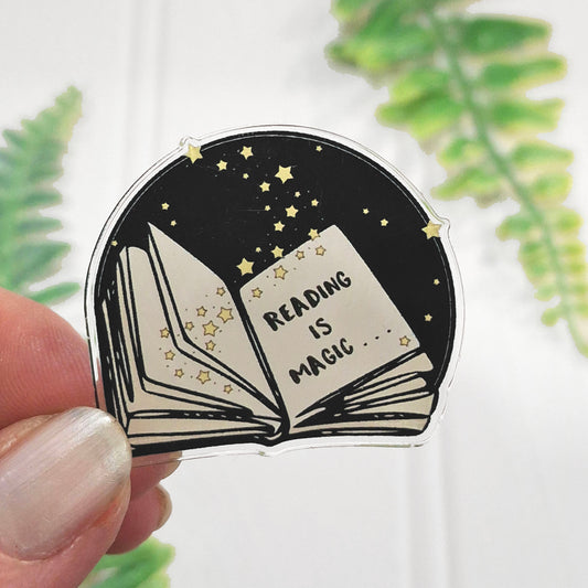 A hand holds an acrylic pin badge featuring an open book with the words 'READING IS MAGIC' on a black background with yellow stars.