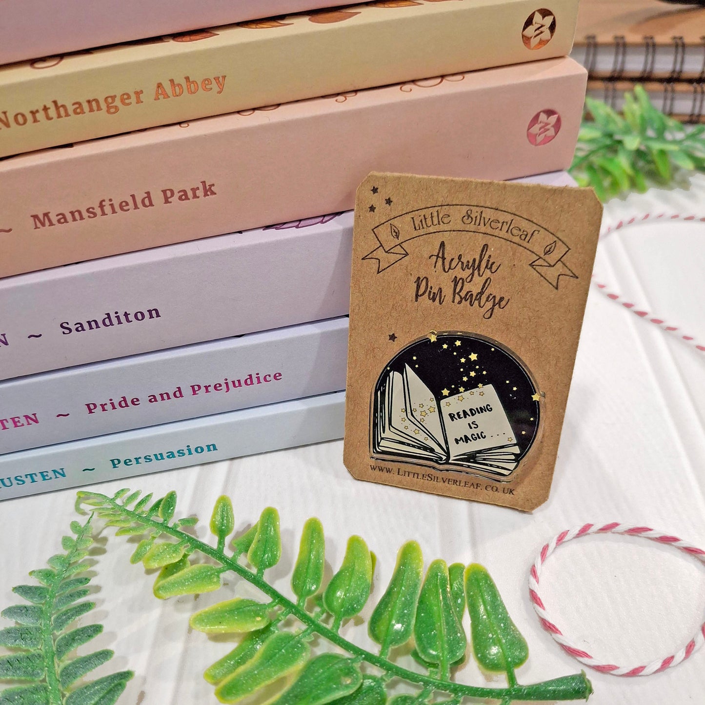 A small round acrylic pin badge on a kraft backing card, styled on a white tabletop with stacked books, green leaves, and a pink-and-white string.