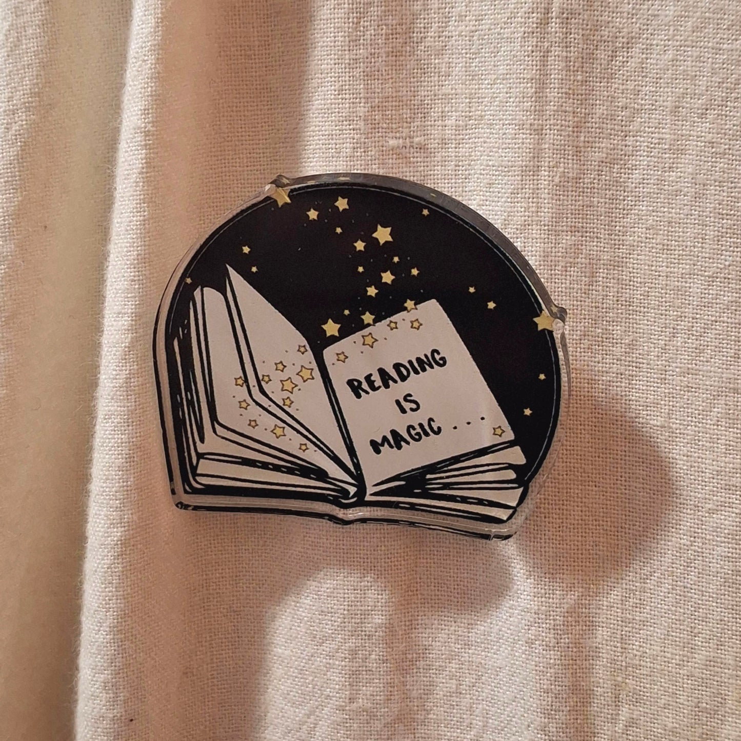 An acrylic pin badge shaped like an open book, featuring a black background with yellow stars and the words 'READING IS MAGIC'.