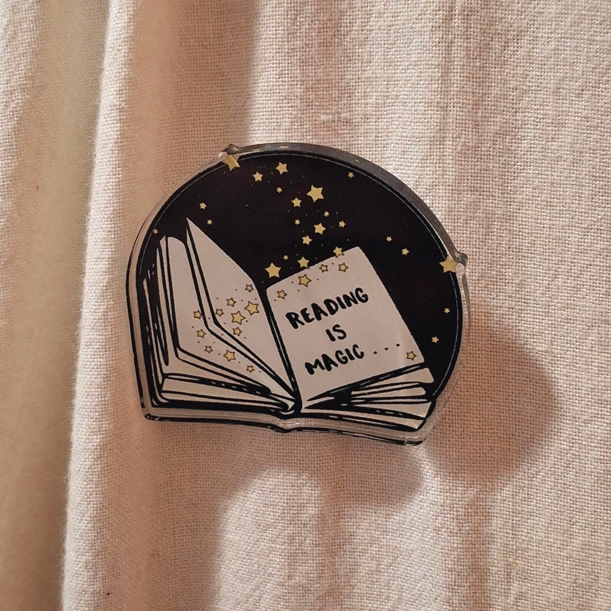 An acrylic pin badge shaped like an open book, featuring a black background with yellow stars and the words 'READING IS MAGIC'.