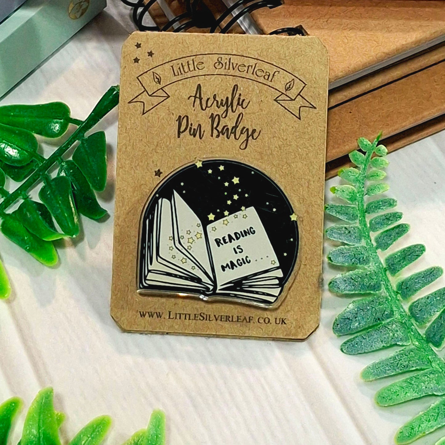 A Little Silverleaf acrylic pin badge on a kraft backing card, featuring an illustration of an open book with the words 'READING IS MAGIC' inside a black starry night sky. The card is styled with green leaves on a white wooden surface.