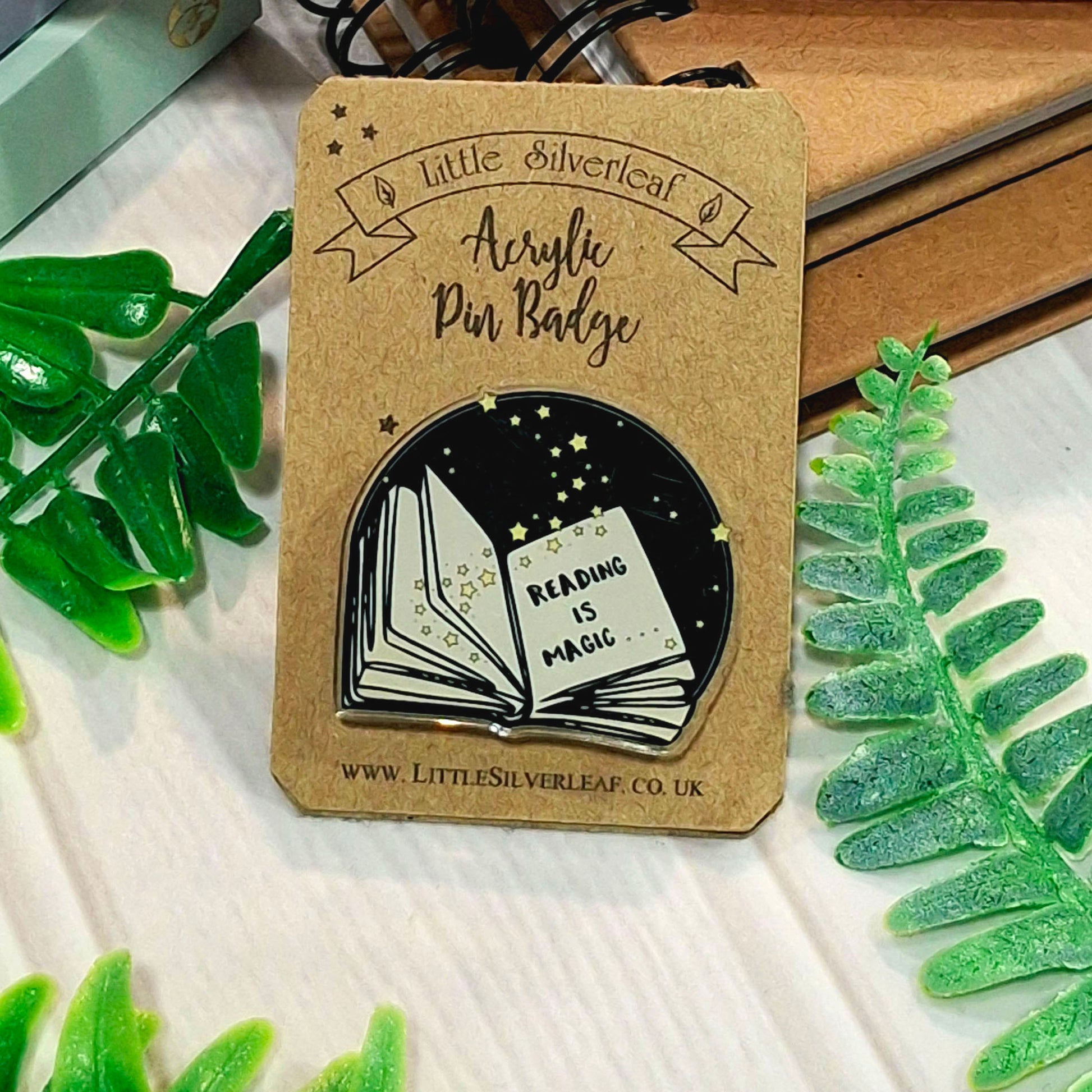 A Little Silverleaf acrylic pin badge on a kraft backing card, featuring an illustration of an open book with the words 'READING IS MAGIC' inside a black starry night sky. The card is styled with green leaves on a white wooden surface.