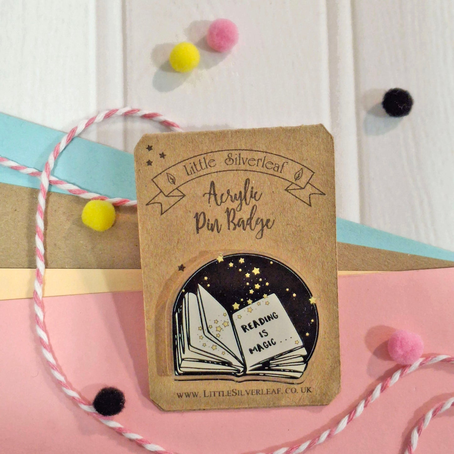 A small acrylic pin badge from Little Silverleaf featuring an open book with the words 'READING IS MAGIC' on a kraft backing card, styled with pastel ribbon, pom-poms, and paper strips.