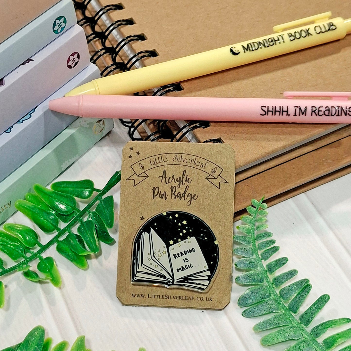 A kraft backing card holding an acrylic pin badge featuring a black illustration of an open book. The badge features the words 'Reading is magic' and the backing card features text 'Little Silverleaf Acrylic Pin Badge' at the top. The card is styled on a white surface with green leaves, notebooks, and two pens, one yellow and one pink.