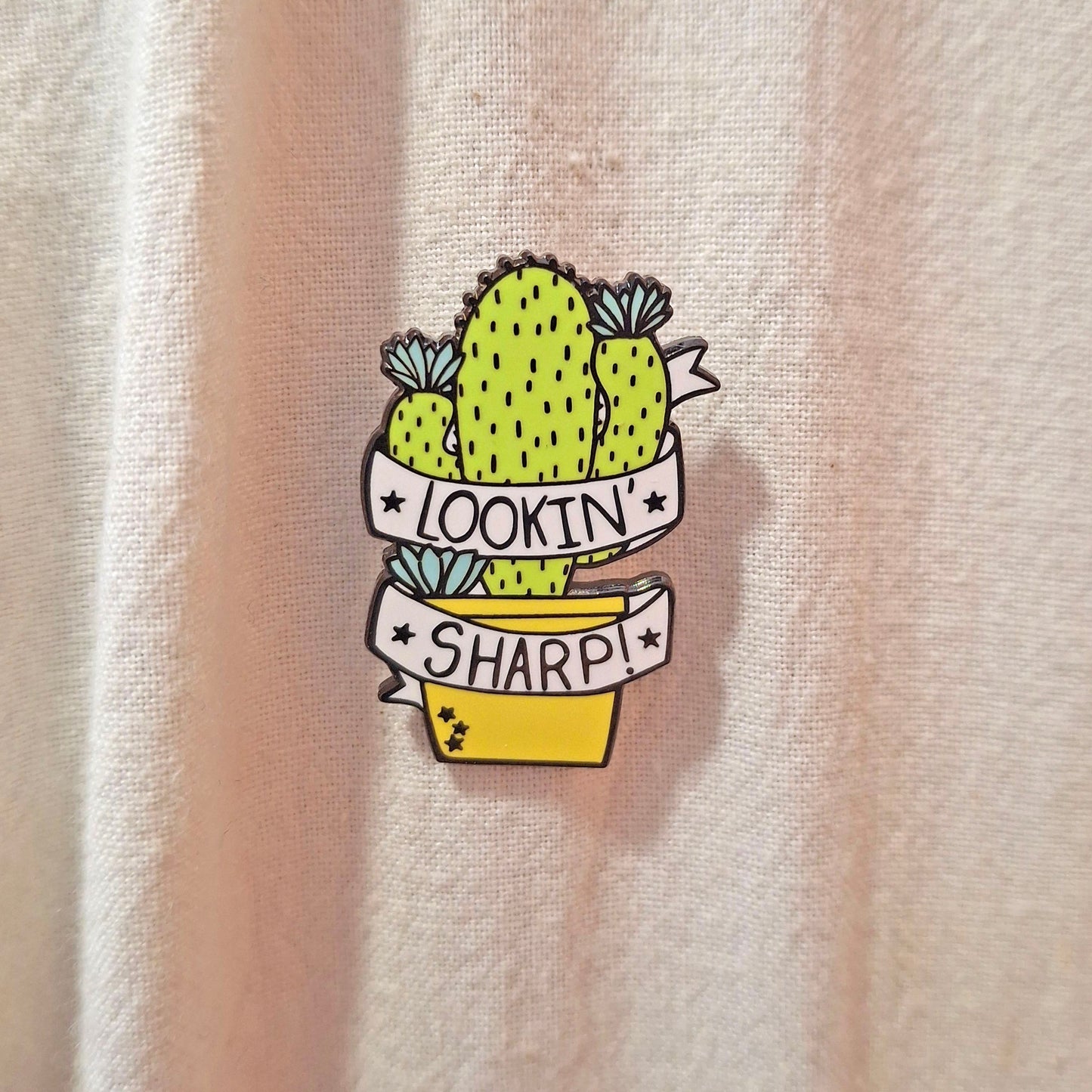 A small enamel pin shaped like a cactus in a yellow pot with white banners reading 'LOOKIN' SHARP!' is shown pinned to a light beige tote bag background.