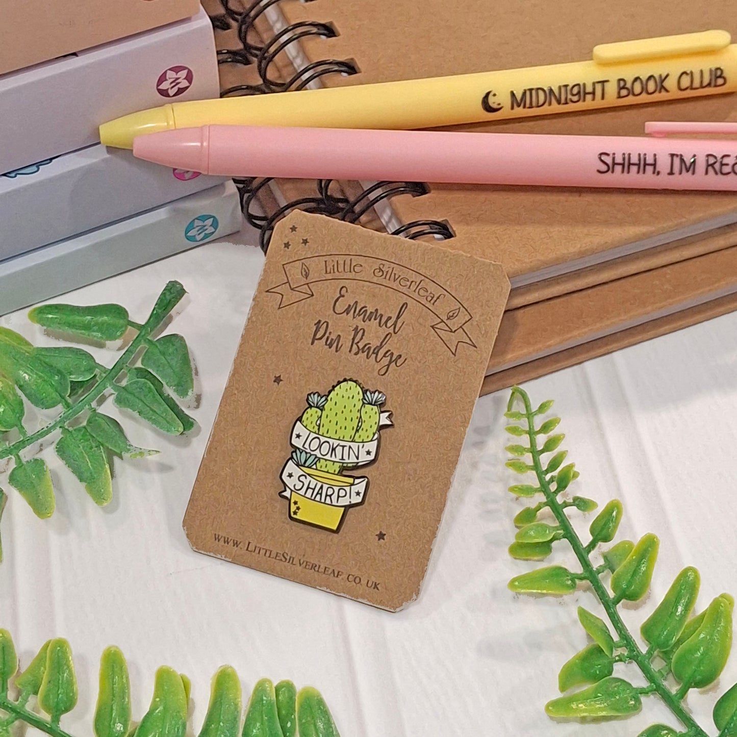 A small enamel pin badge displayed on a kraft backing card, styled with notebooks, pens, and green leaves on a white surface. The pin features a cactus holding a sign that reads 'Lookin' Sharp!'.