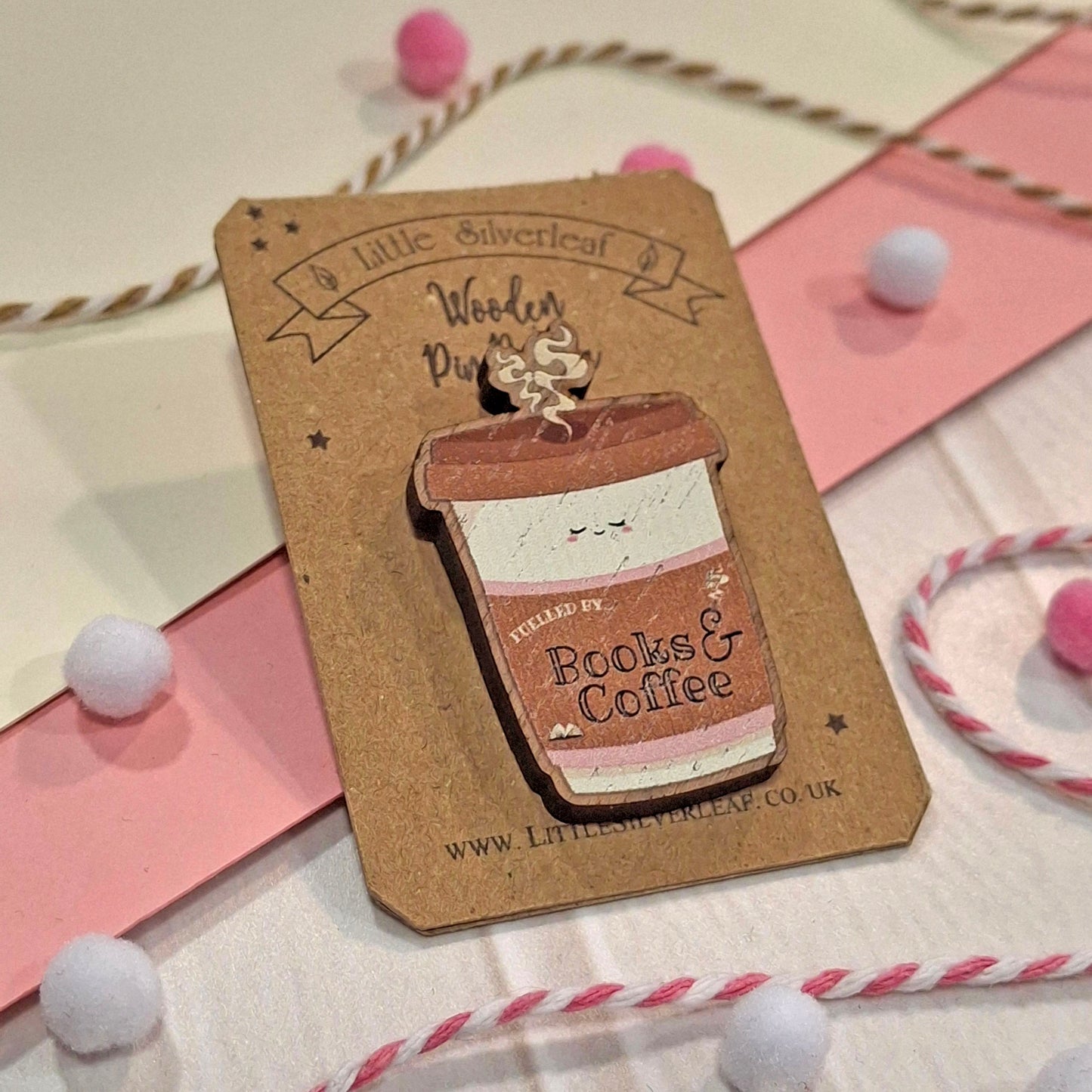 A cute wooden pin shaped like a coffee cup with a smiling face, mounted on a brown kraft card. The card is branded 'Little Silverleaf Wooden Pin Badge' and is styled on a white surface with pink paper strips, white pom-poms, and twisted twine.