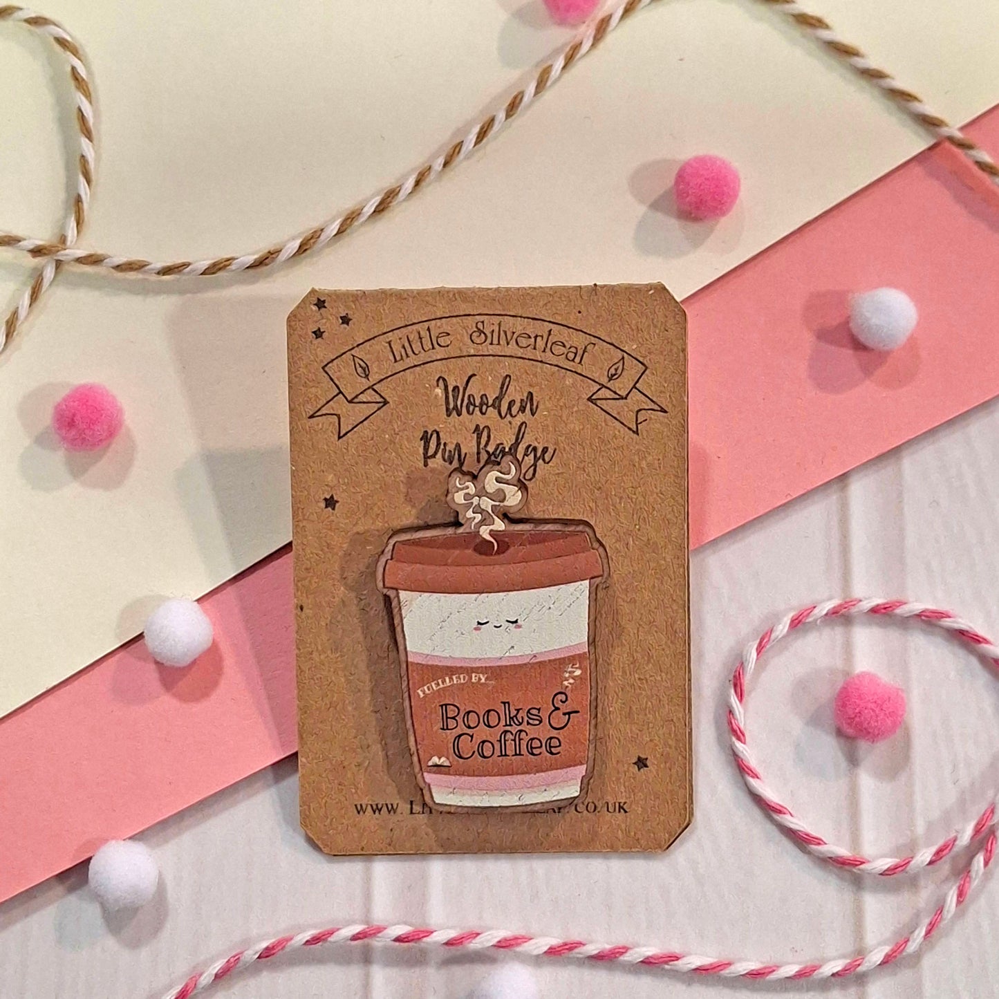 A wooden pin badge shaped like a coffee cup, attached to a brown kraft card labelled 'Little Silverleaf Wooden Pin Badge'. The badge is styled on a white surface with pink and cream paper strips, bakers twine, and small pink and white pom-poms.