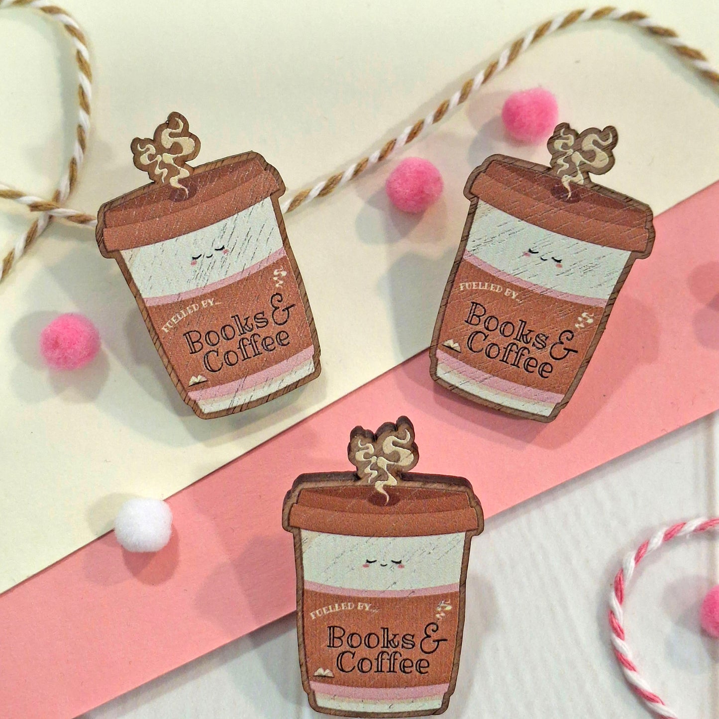 Three coffee cup shaped pin badges with the words 'FUELLED BY Books & Coffee' are arranged on a cream and pink background with string and pom-pom decorations.