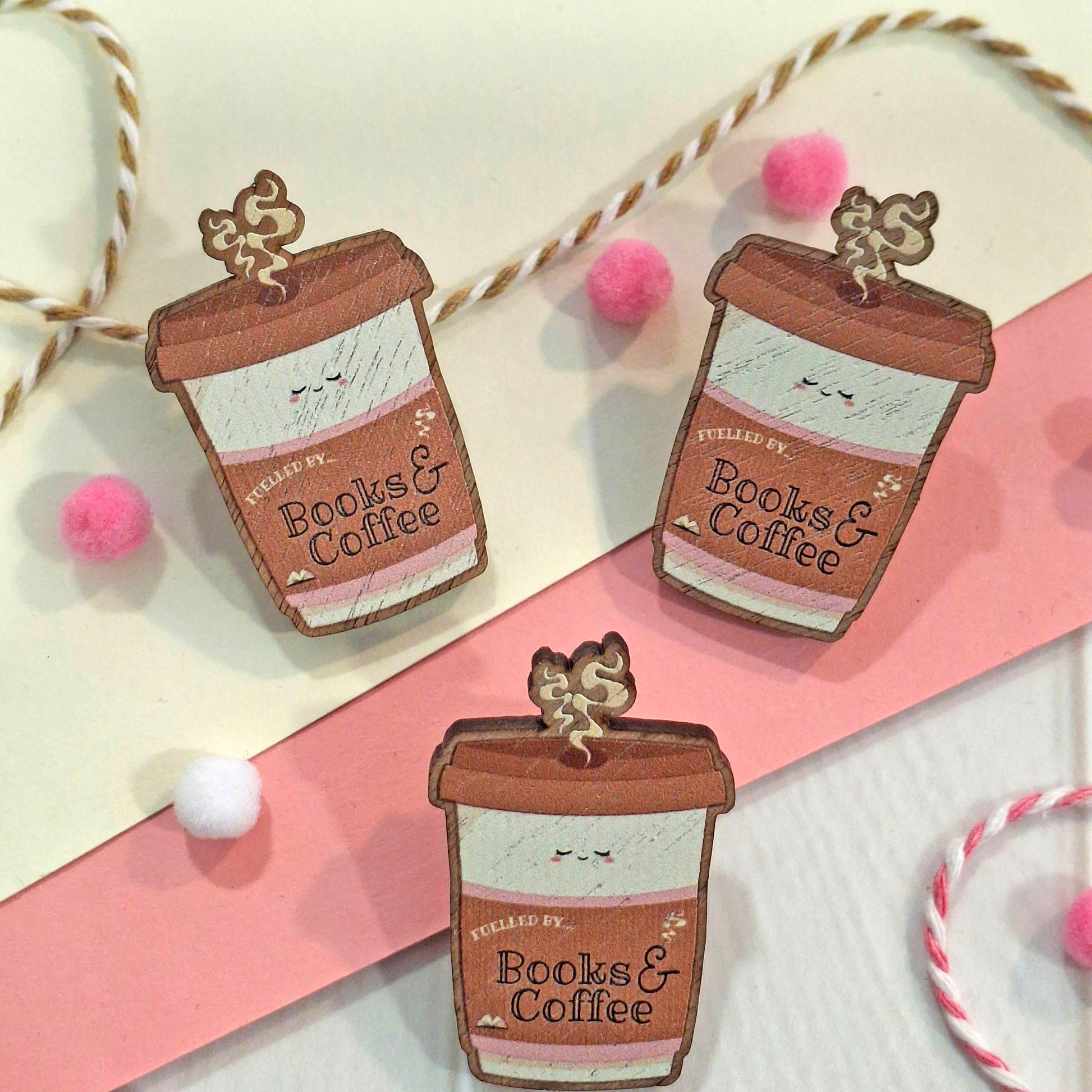 Three coffee cup shaped pin badges with the words 'FUELLED BY Books & Coffee' are arranged on a cream and pink background with string and pom-pom decorations.