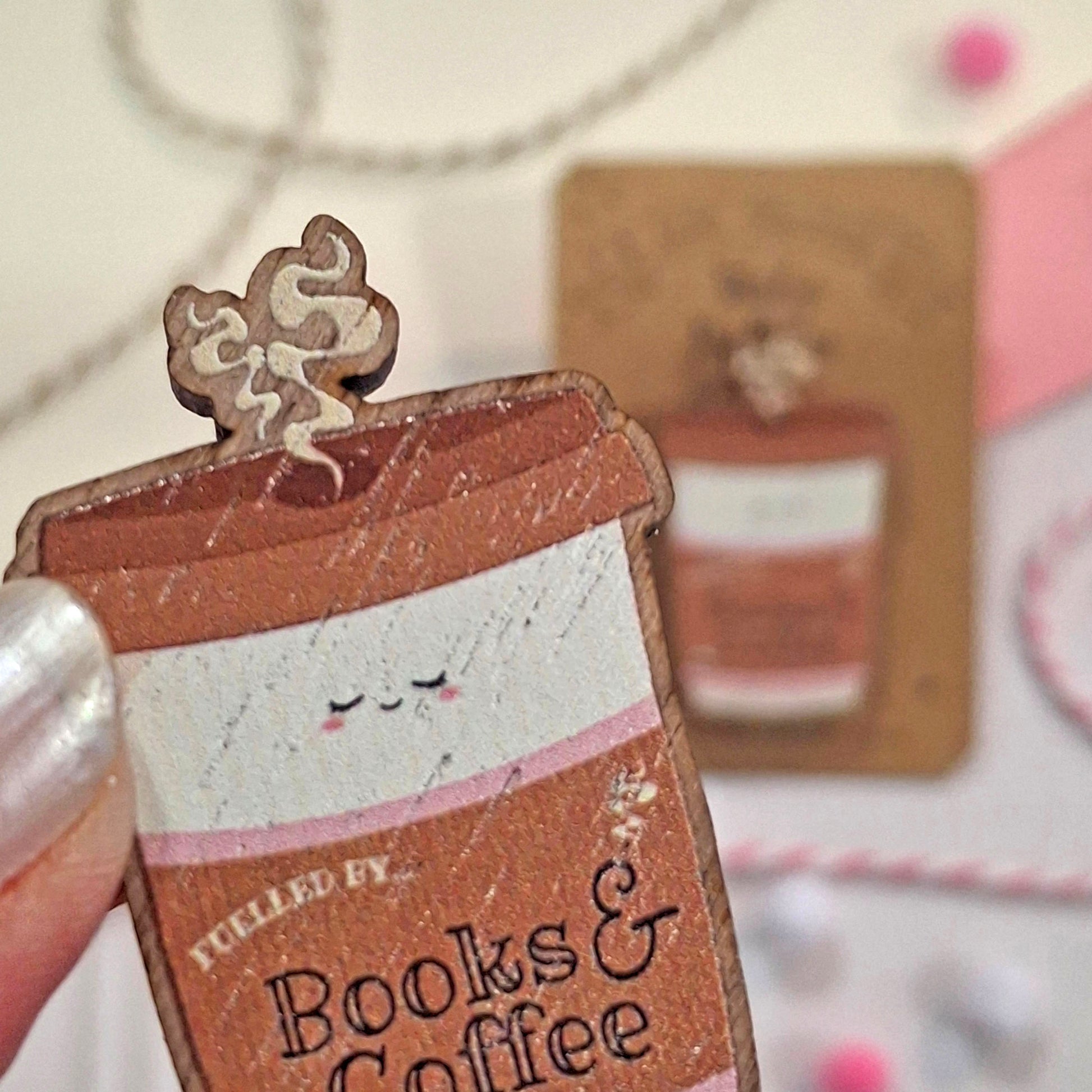 A close-up of a hand holding a small illustrated coffee cup wooden pi badge with the text 'FUELLED BY Books & Coffee' and a decorative lid. The background is softly blurred with similar items and pastel tones.