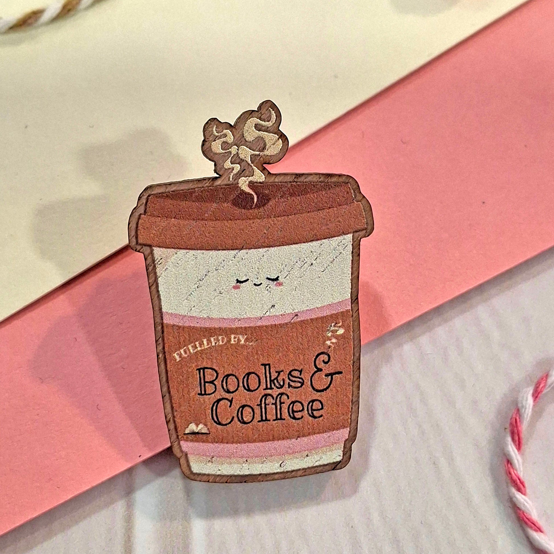 A cute kawaii coffee cup wooden pin with the text 'FUELLED BY Books & Coffee' and a smiling face, resting on a pink card over a white surface.