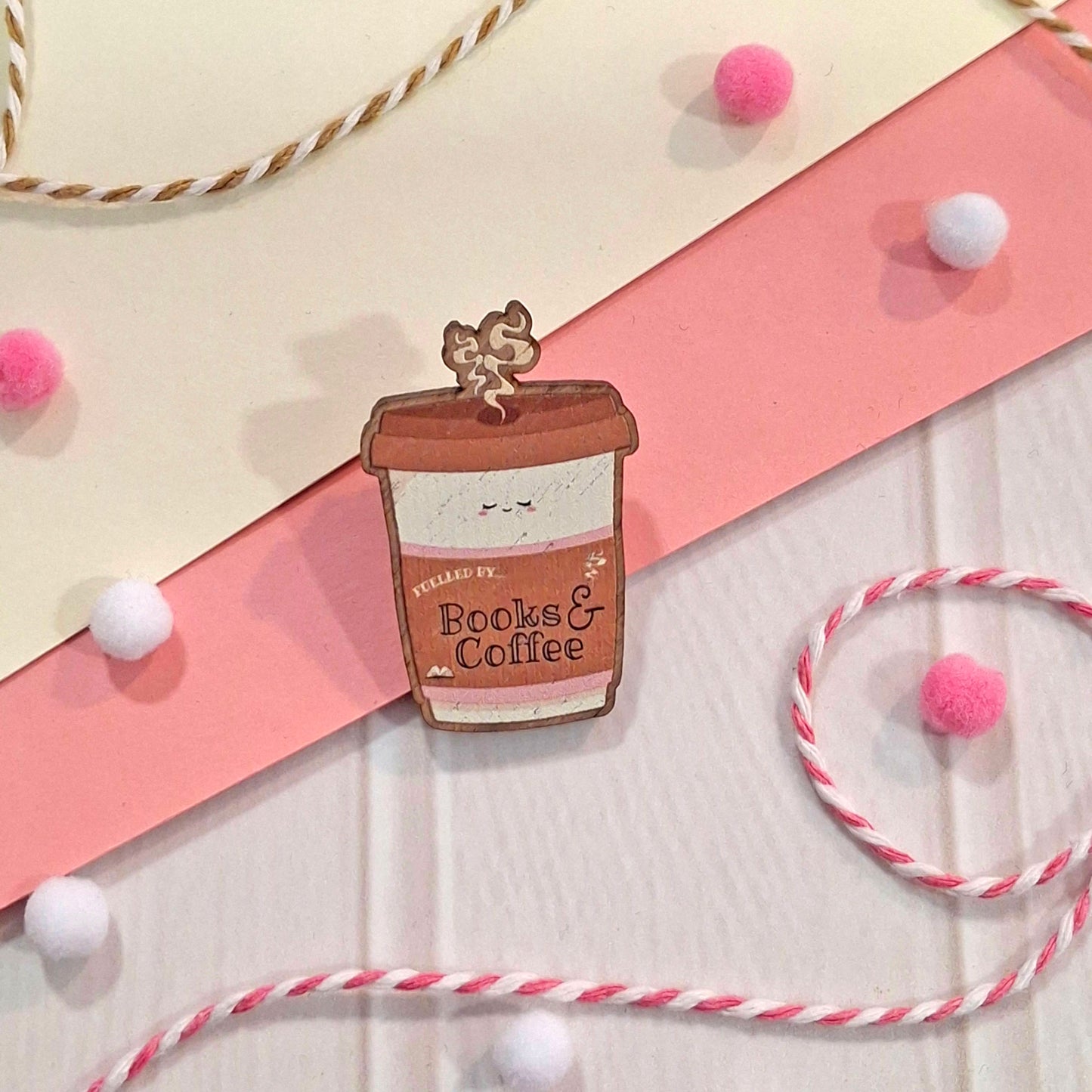 A cute wooden pin shaped like a coffee cup with a smiling face and the text 'Fuelled by Books & Coffee', displayed on a styled flat-lay background with pink and cream paper, white and pink pom-poms, and twisted twine.