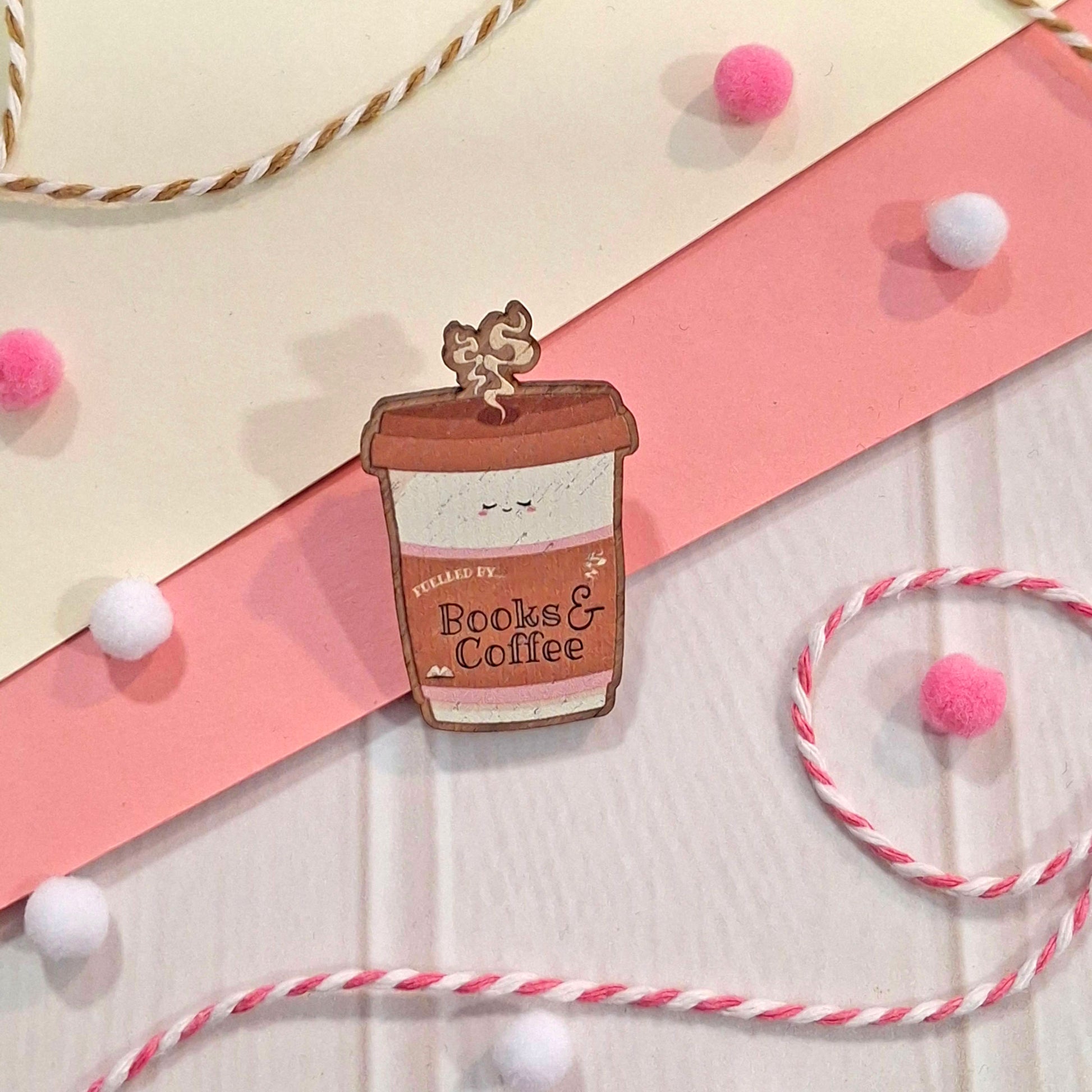 A cute wooden pin shaped like a coffee cup with a smiling face and the text 'Fuelled by Books & Coffee', displayed on a styled flat-lay background with pink and cream paper, white and pink pom-poms, and twisted twine.