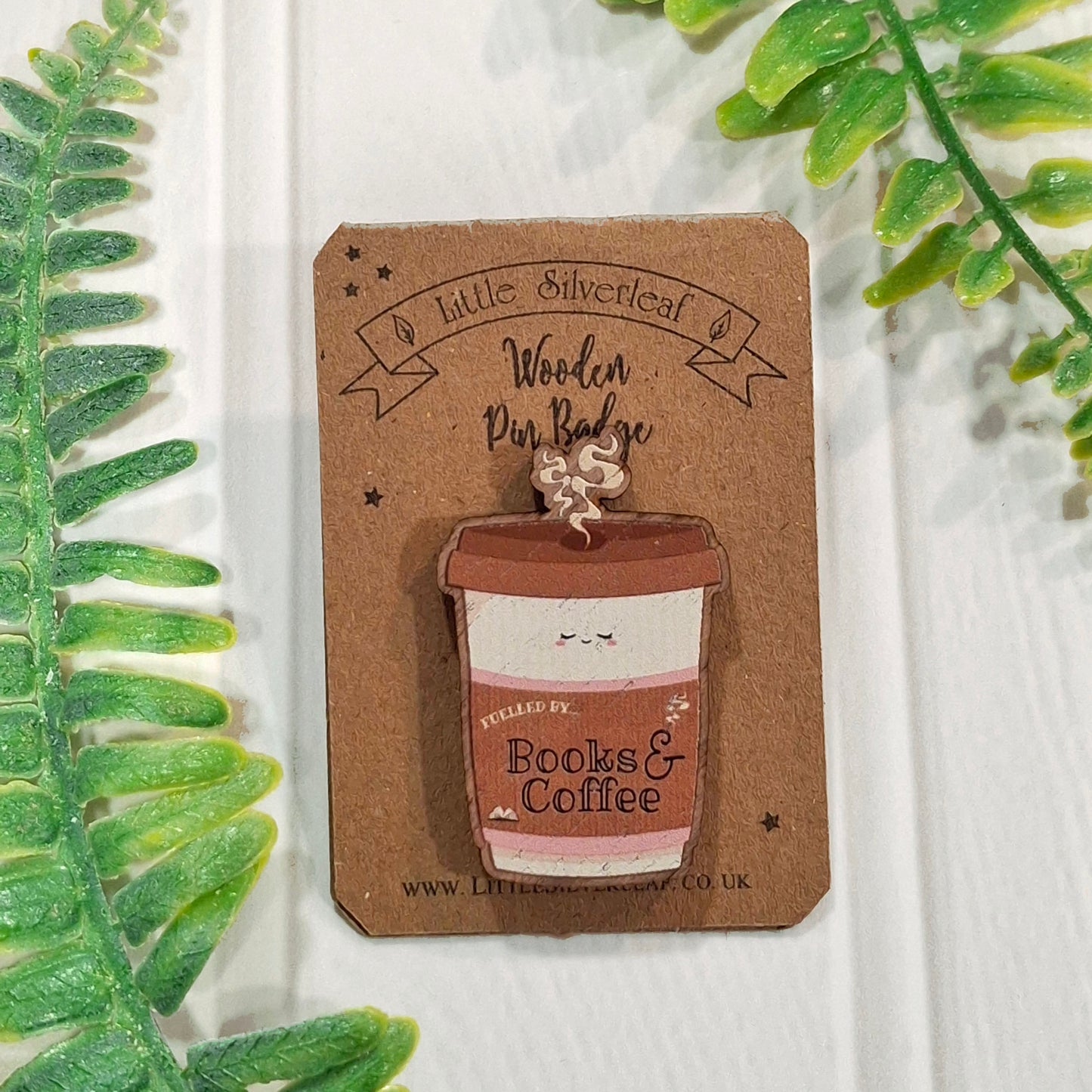 A wooden pin shaped like a coffee cup, attached to a brown cardboard display card. The card is photographed on a white wood surface with green leaves framing the edges.