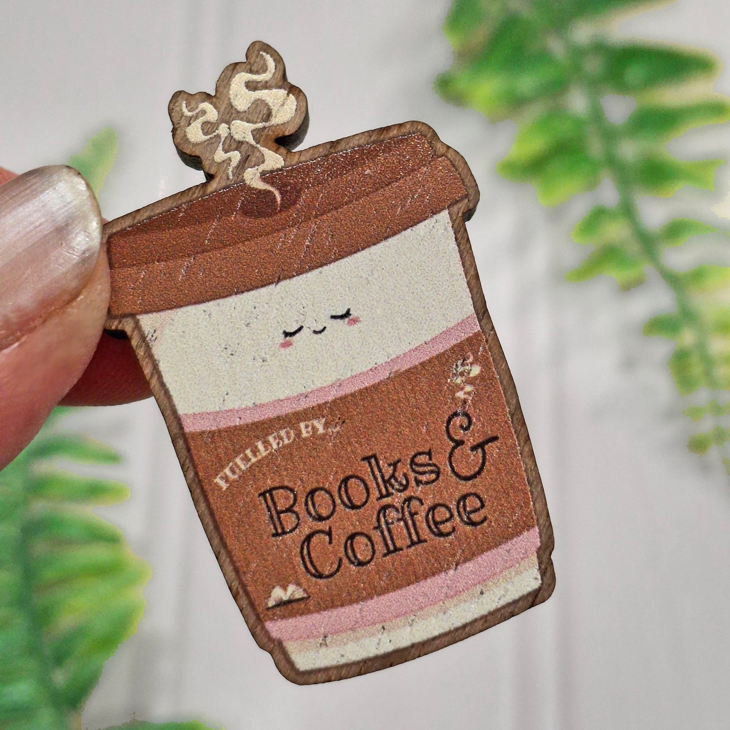 A close-up of a hand holding a small coffee cup-shaped wooden pin badge that reads 'Fuelled by Books & Coffee.' The item has a brown and cream design with pink stripes and a small decorative swirl on top, against a softly blurred background with green leaves.